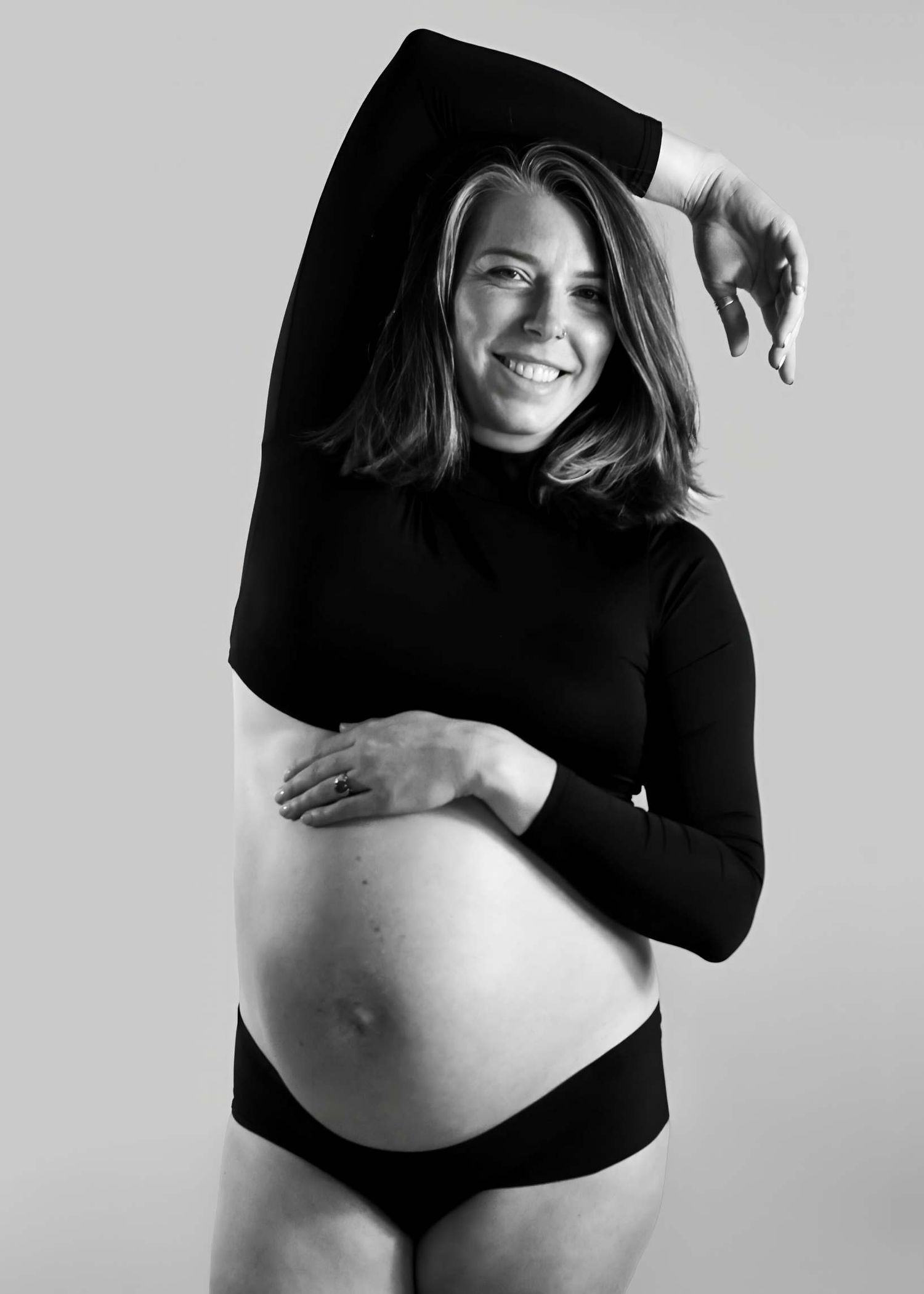 Black and white maternity photo of smiling pregnant woman in black turtleneck top posing with hand raised above head. Redmond Oregon studio
