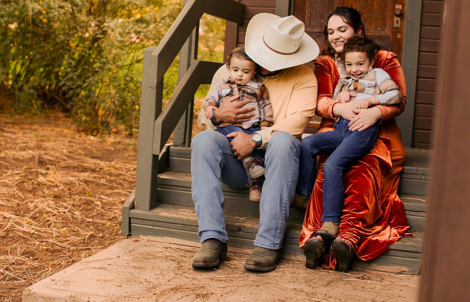 Family with two babies sitting on cabin porch steps, man in cowboy hat, woman in red dress, fall foliage background.