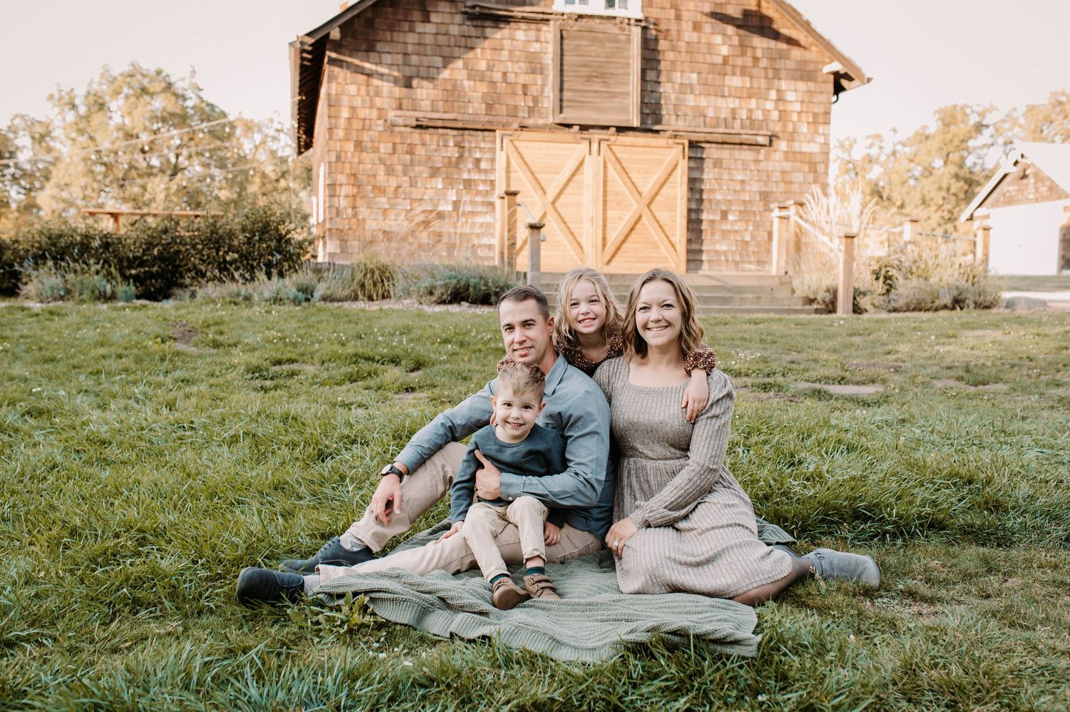 Happy family of four posing on a blanket in a green field with a rustic barn in the background.