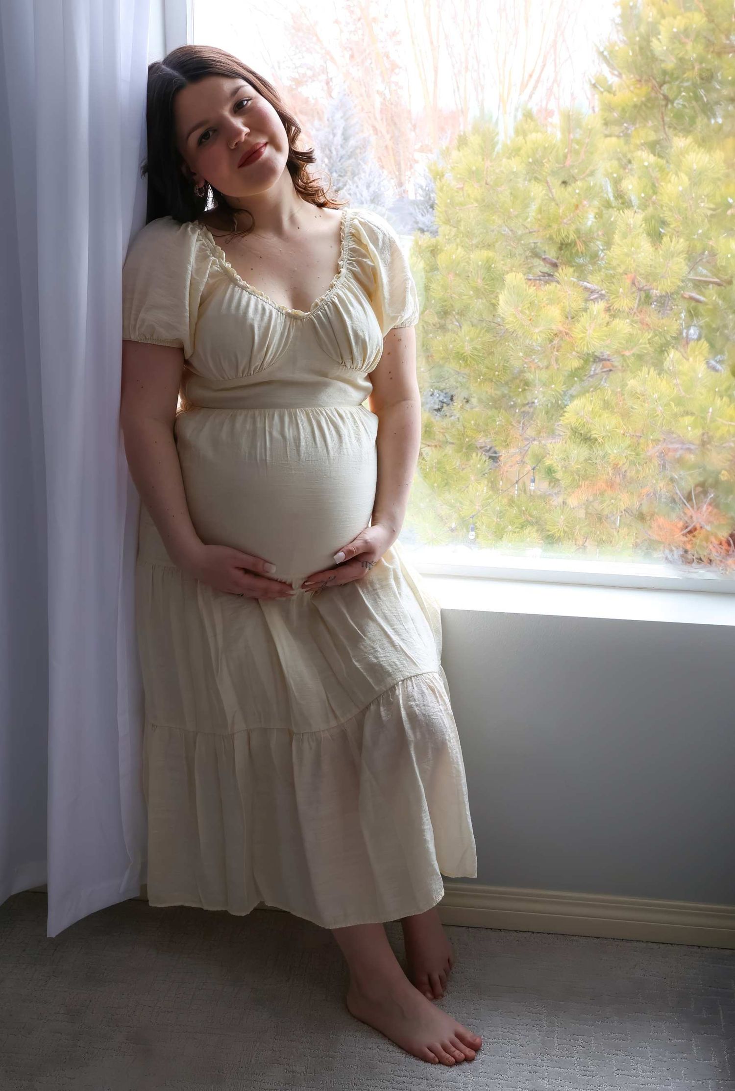 Smiling pregnant woman in cream dress cradling baby bump, standing barefoot by sunlit window with trees outside.