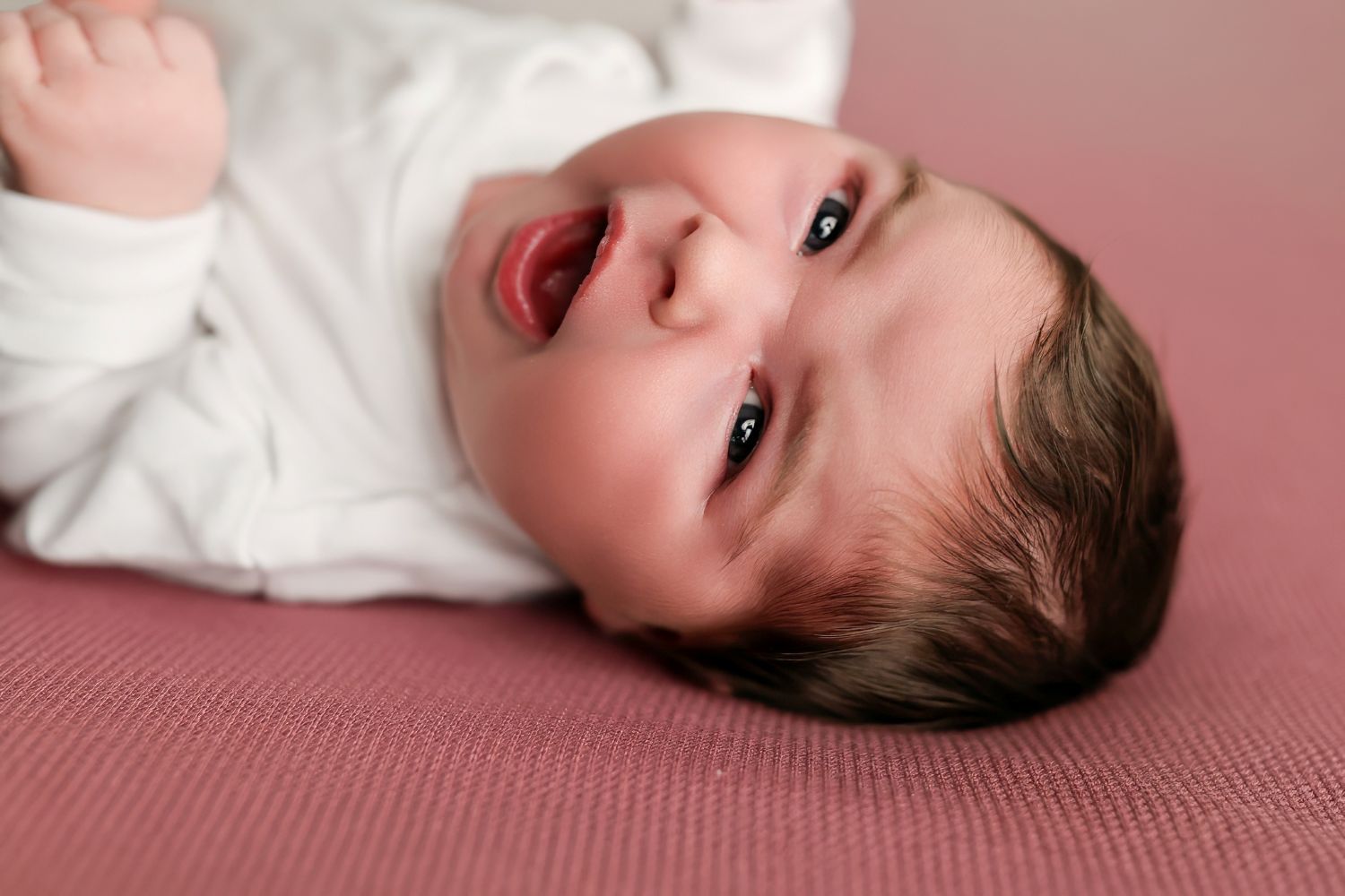 Newborn baby with dark hair lying on pink blanket, eyes open and mouth slightly agape.