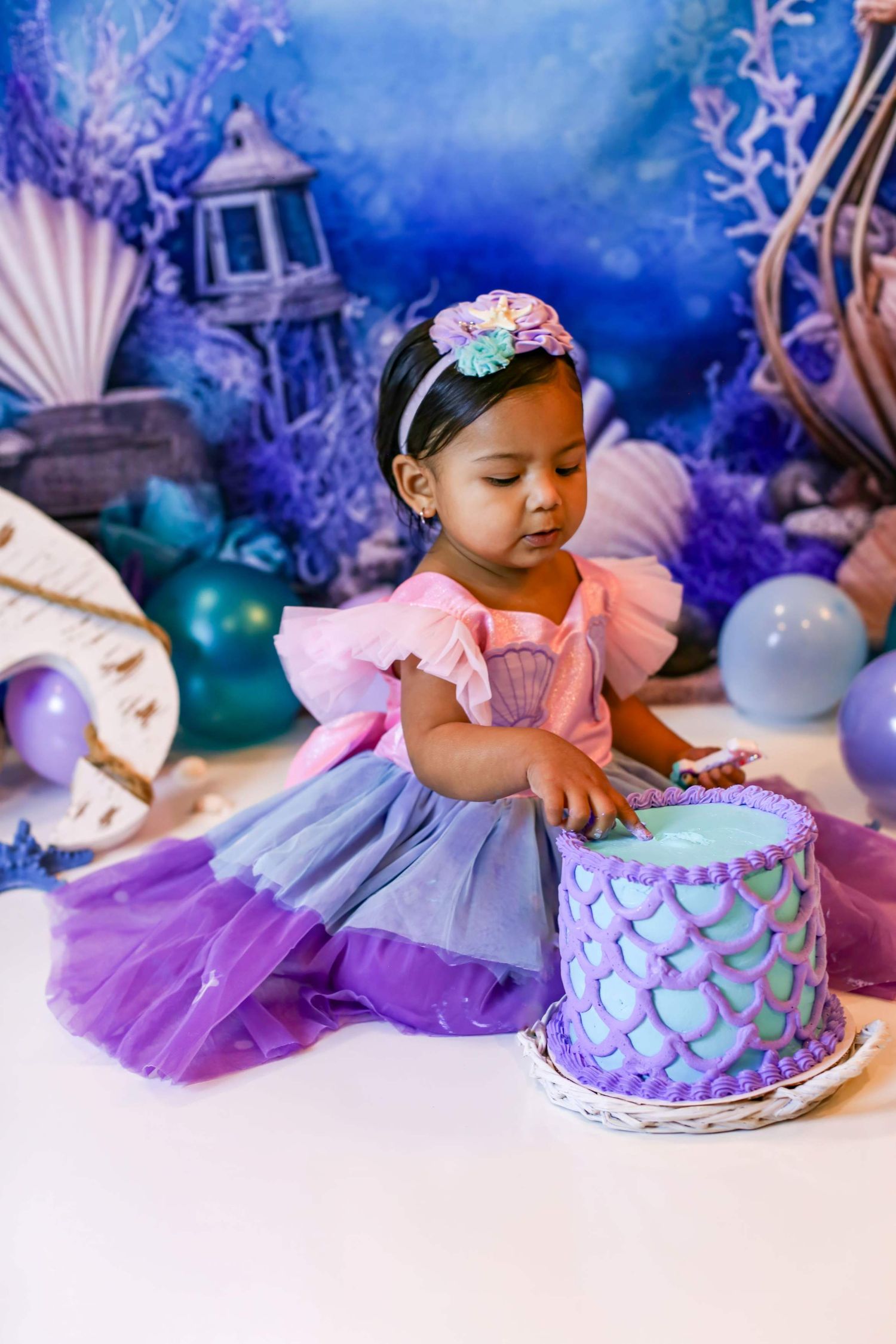 Toddler girl in mermaid dress touching her first birthday cake at an underwater themed party.