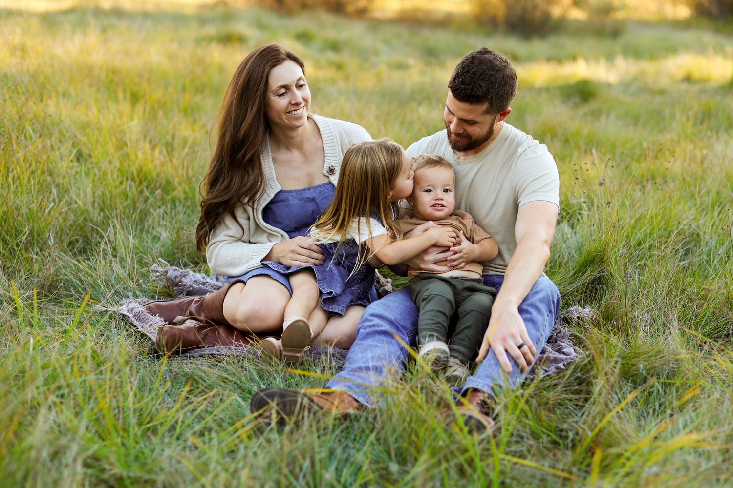 Family of four sitting on a blanket in a green field, parents smiling as children embrace in a warm outdoor moment.