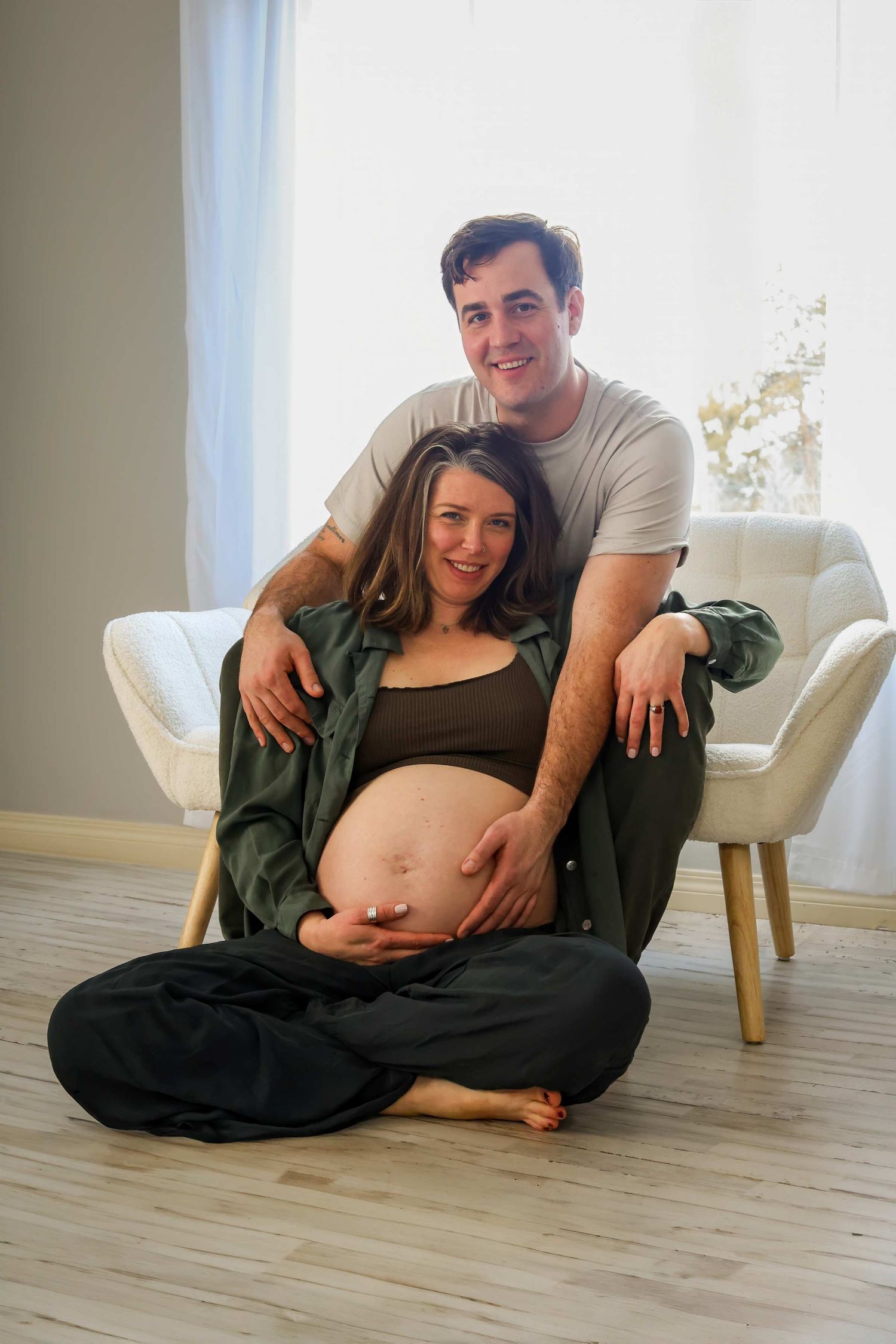 A couple poses together on a hardwood floor during a maternity photoshoot showing the progression of pregnancy.