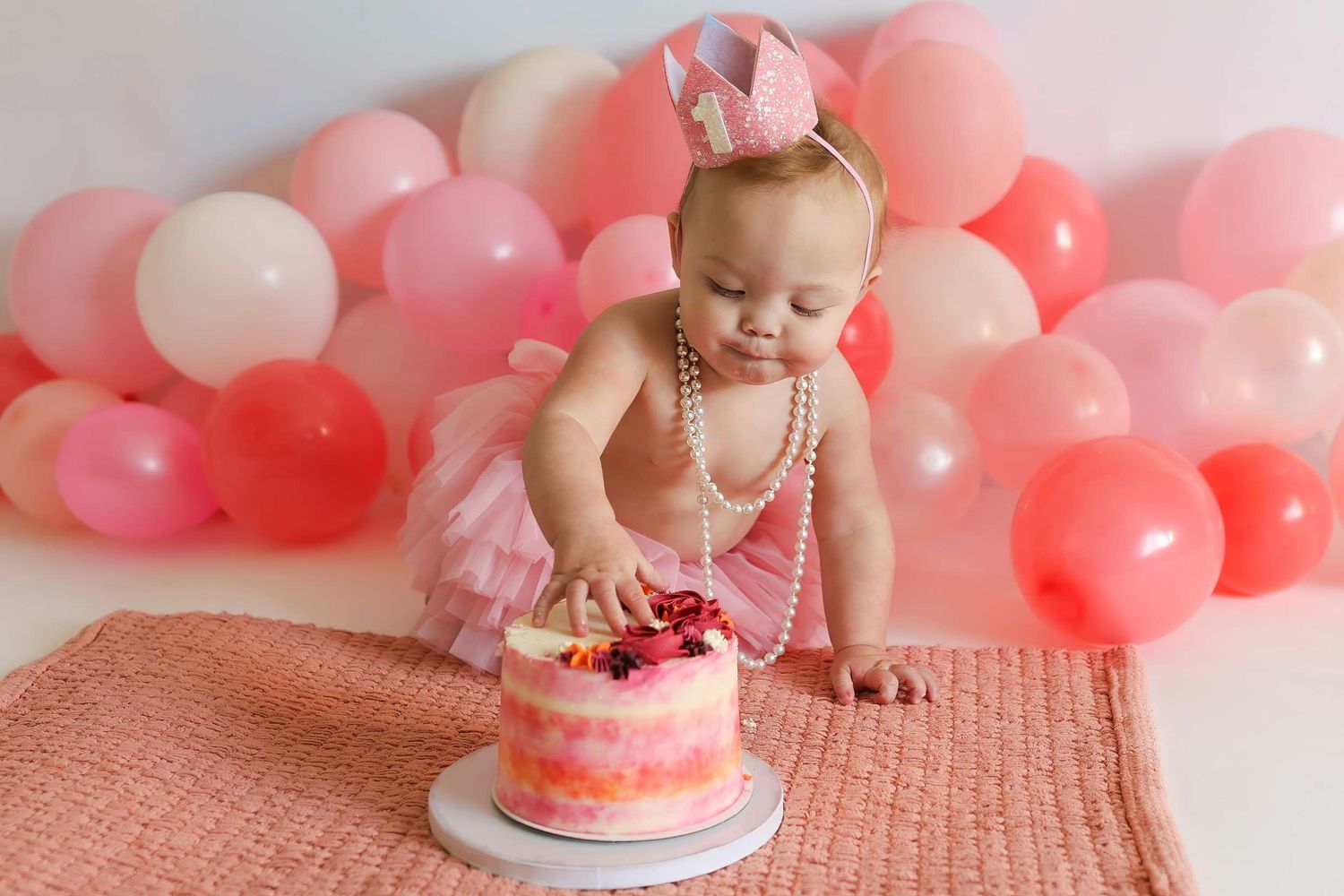 Baby in pink dress celebrates first birthday with smash cake surrounded by pink and coral balloons. Redmond Oregon Studio