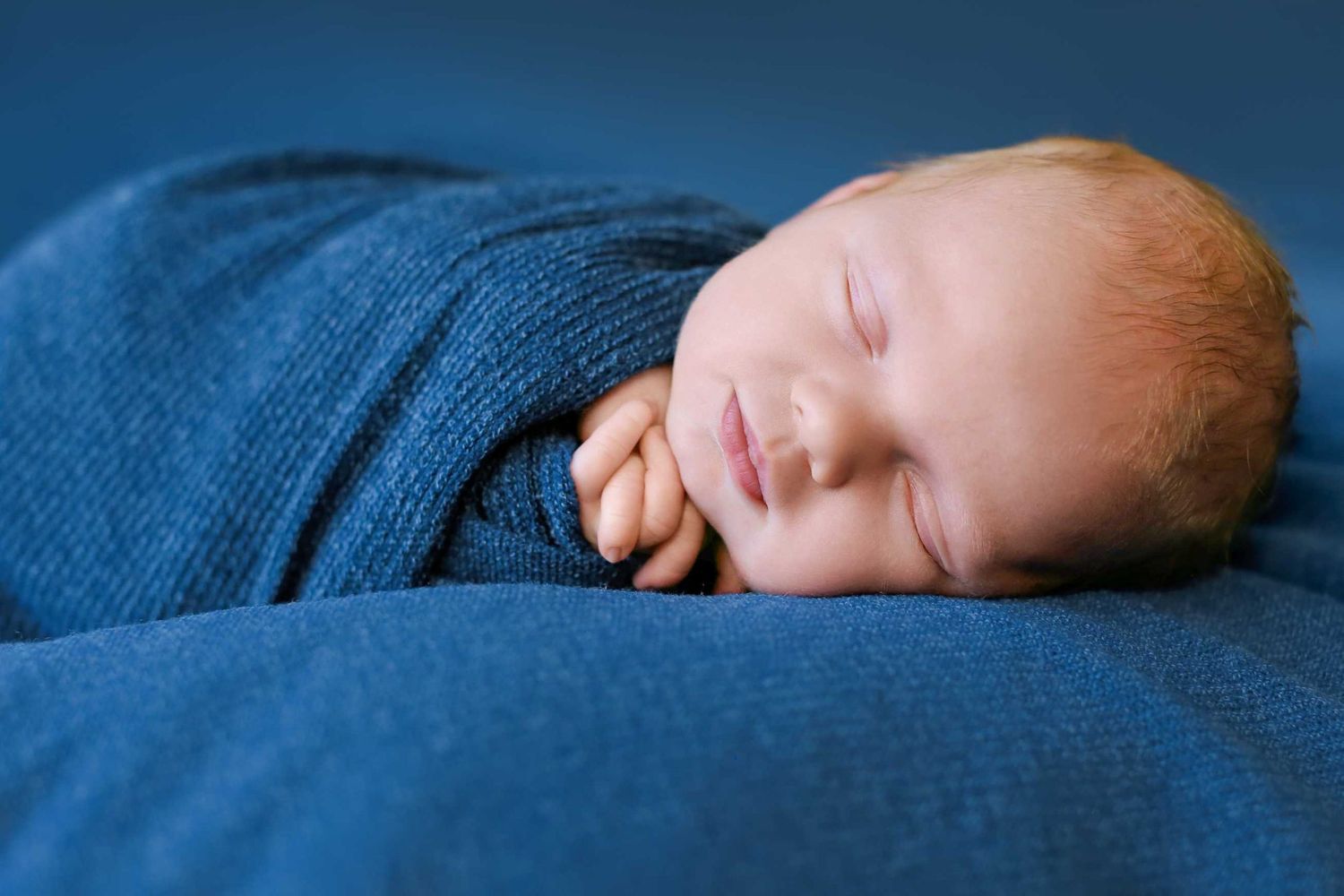 A sleeping newborn baby rests peacefully on blue textured fabric in a studio portrait setting.