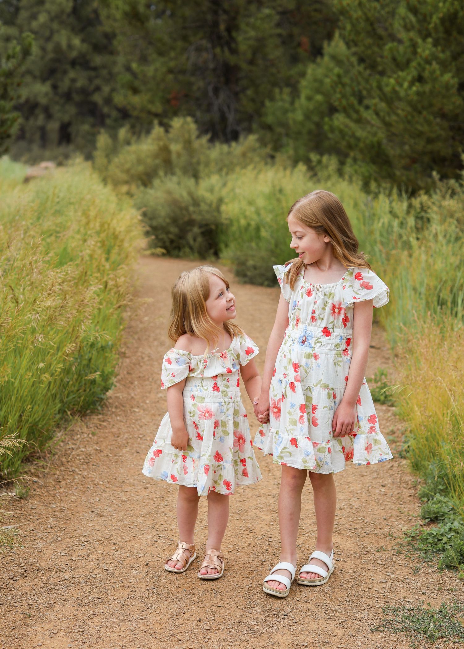Two young girls in matching floral dresses hold hands on a dirt path surrounded by lush greenery.