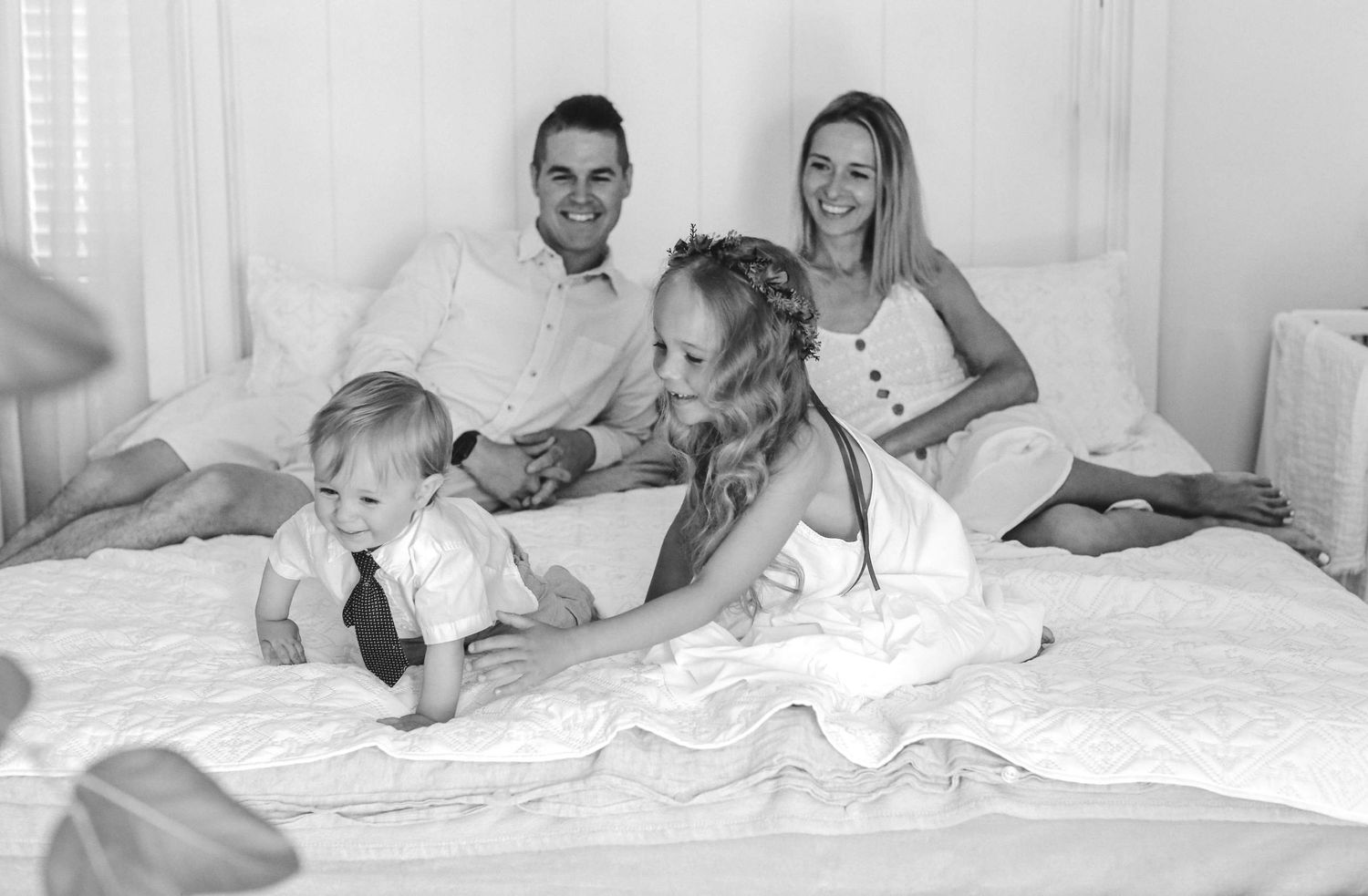 A happy family with a baby boy in a tie posing together on a white bed in a black and white photo.