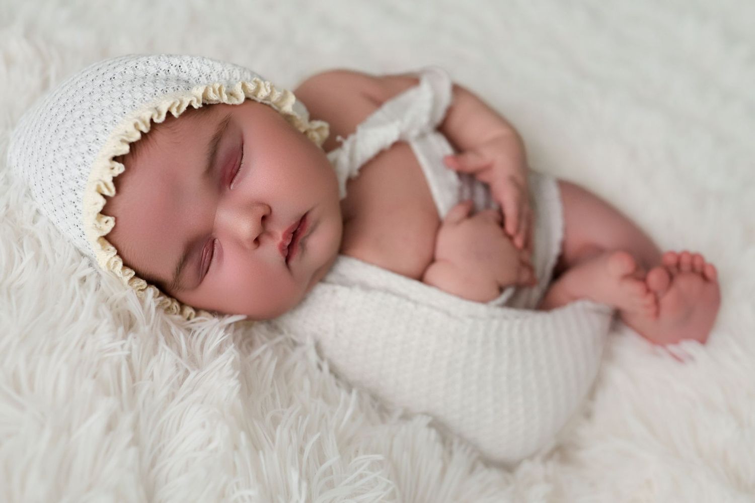 Sleeping newborn wrapped in white knit blanket wearing delicate lace bonnet on soft fur backdrop.