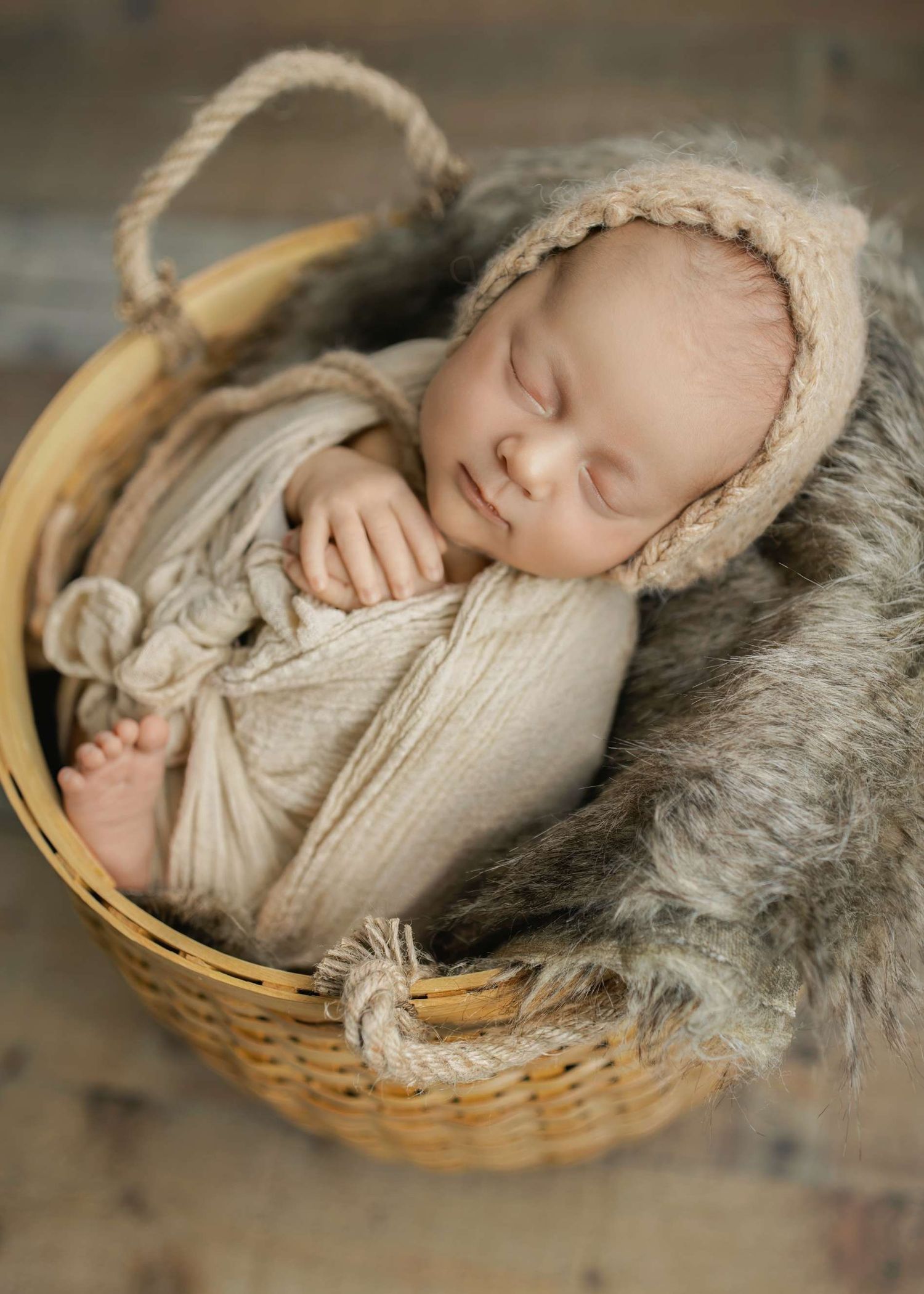 Newborn baby resting in cozy basket with beige bonnet and grey fur blanket.