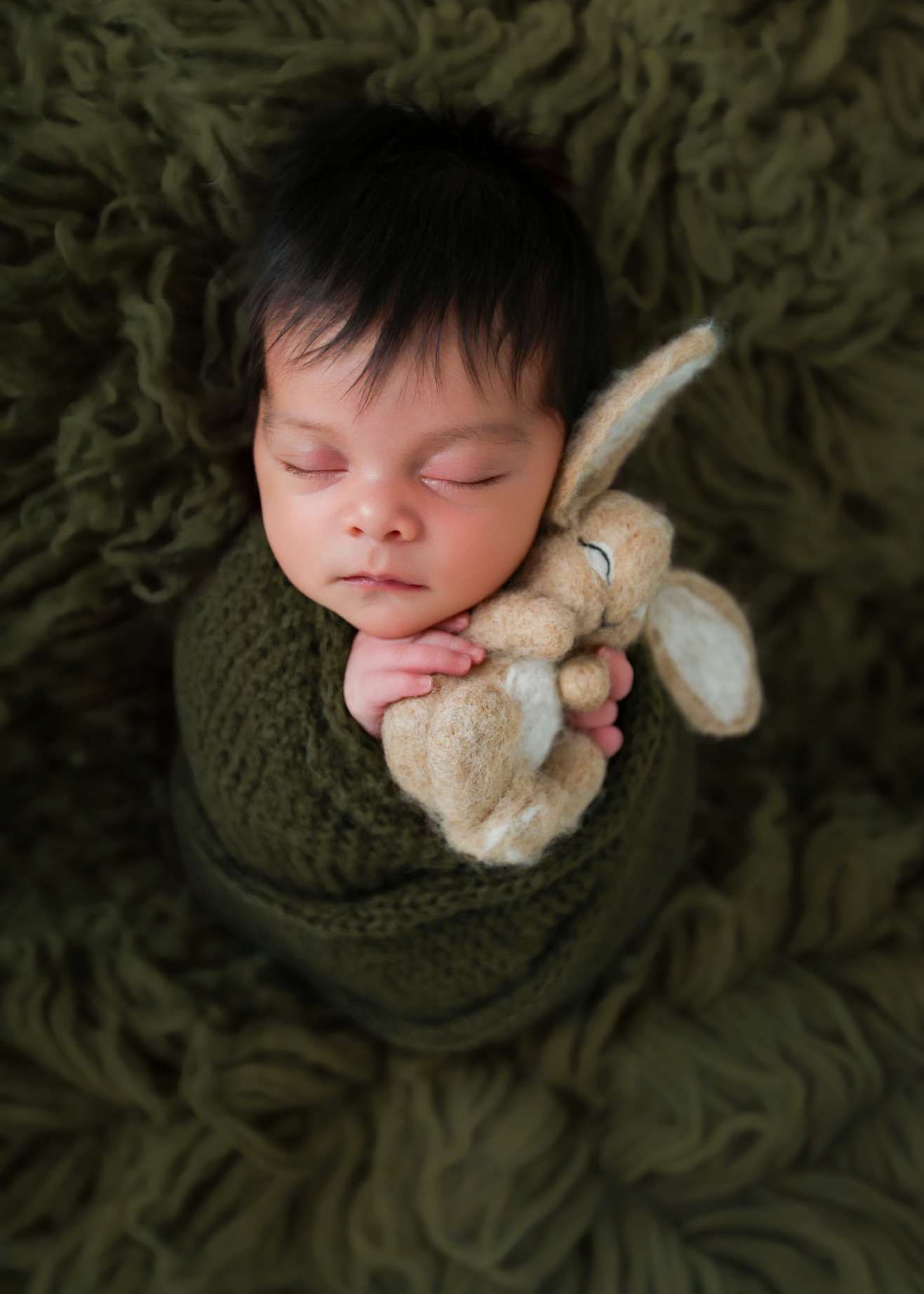 Sleeping newborn swaddled in green knit wrap holding a stuffed bunny on a green flokati rug.