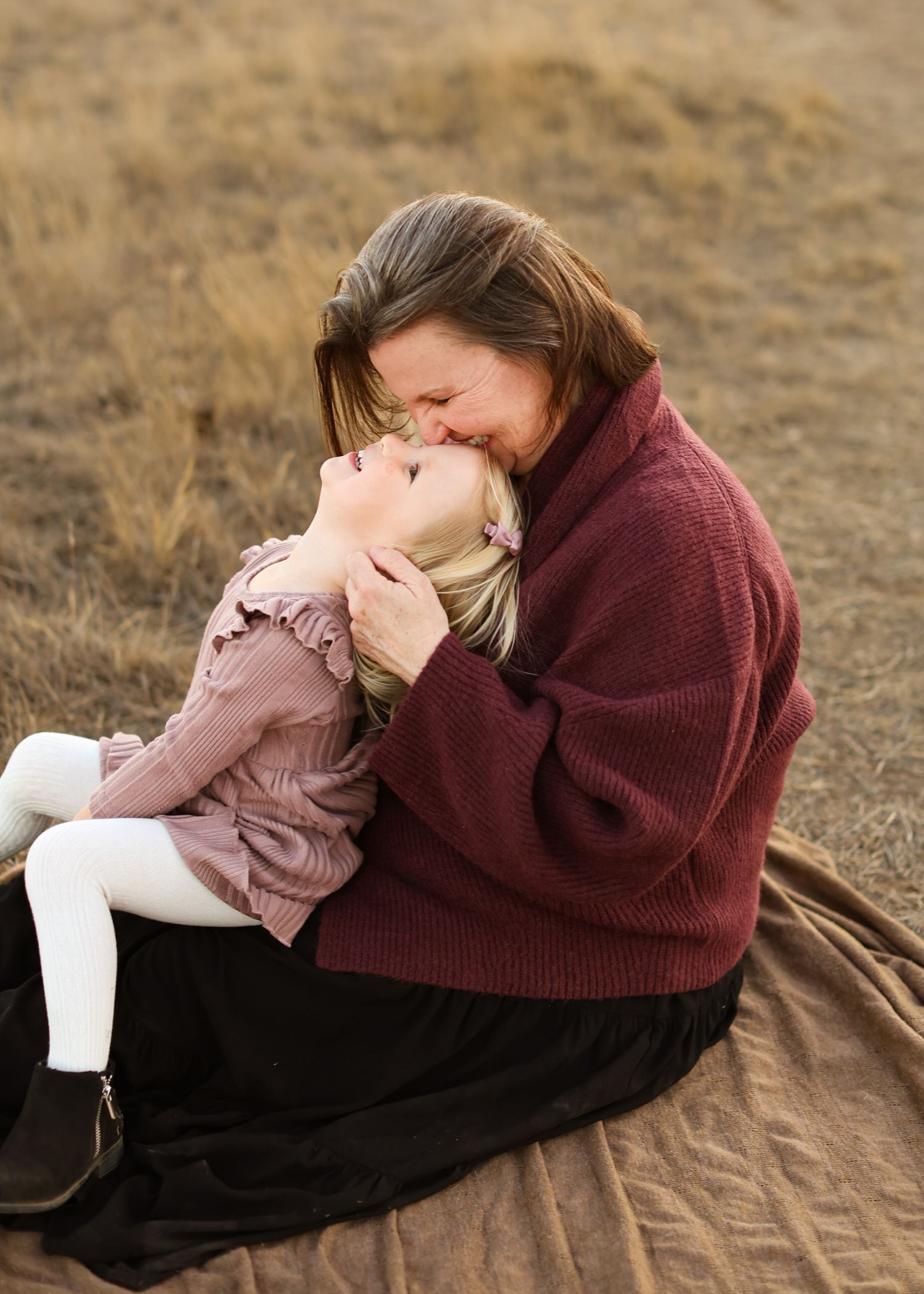 A smiling woman in a burgundy sweater cuddles a laughing young girl while sitting on a tan blanket in a golden grass field.
