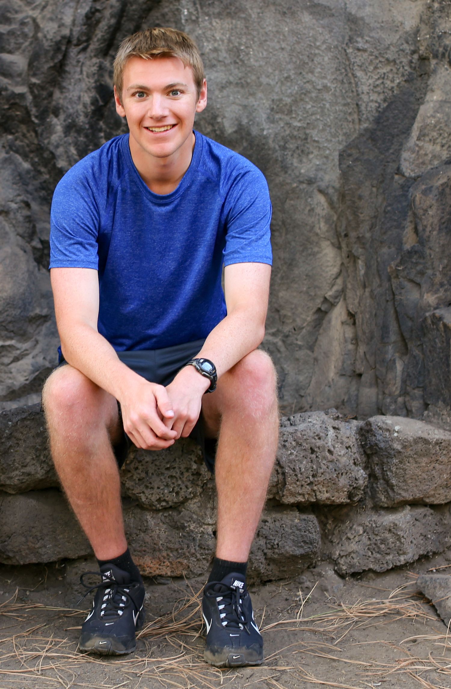 Young man in blue t-shirt and shorts sitting on rocky surface, smiling outdoors.