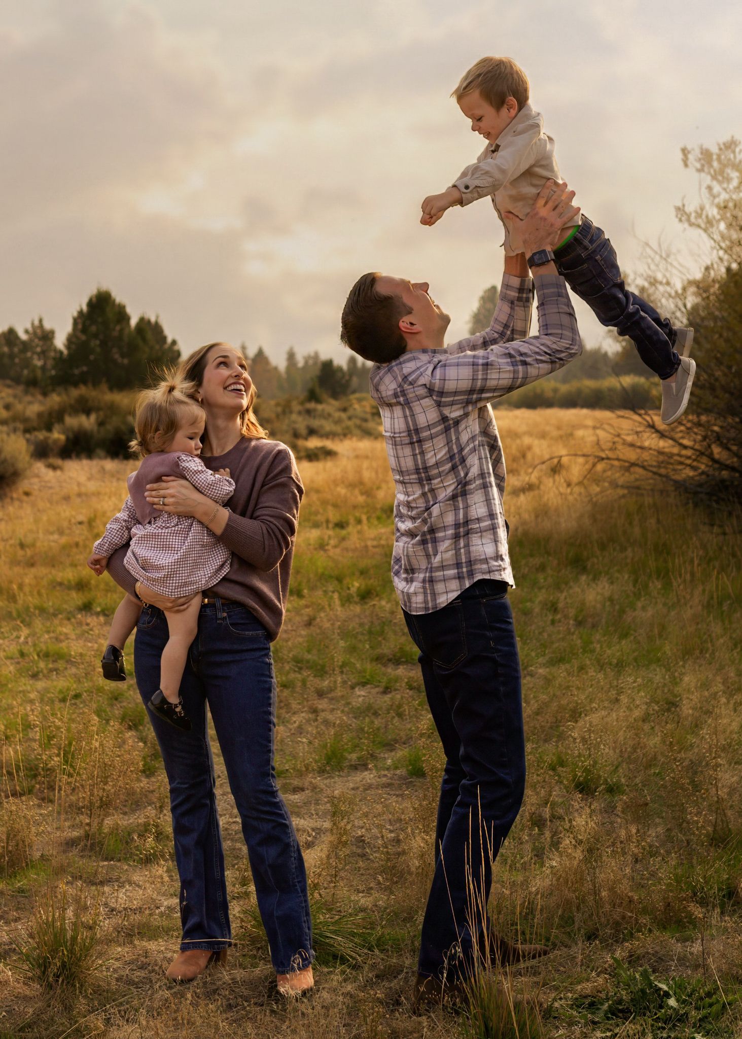 A joyful family of four plays outdoors, with a man lifting a toddler high while a woman holds a young girl in a field.