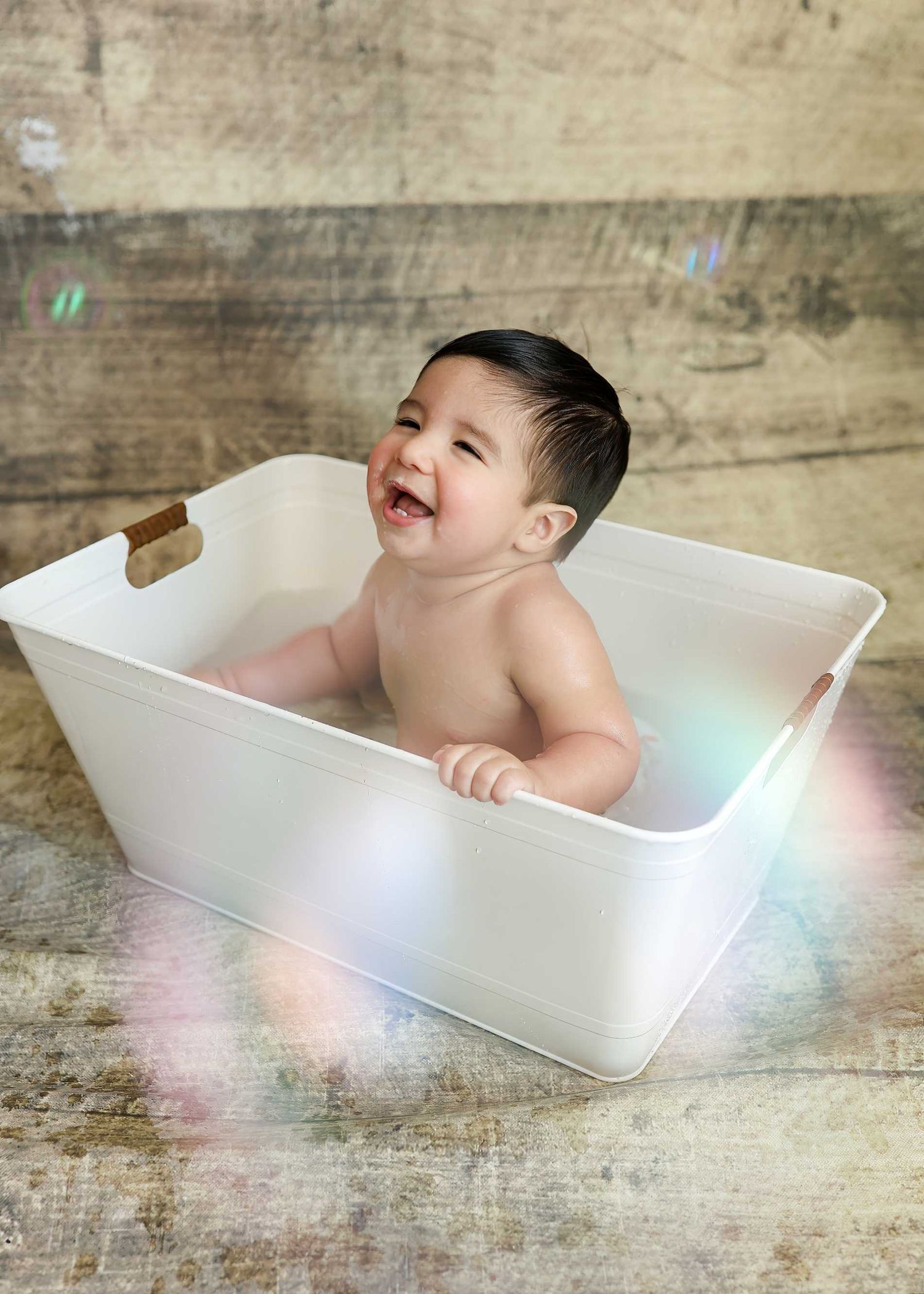 Happy baby laughing in a white metal tub with wooden handles during a professional photo shoot.