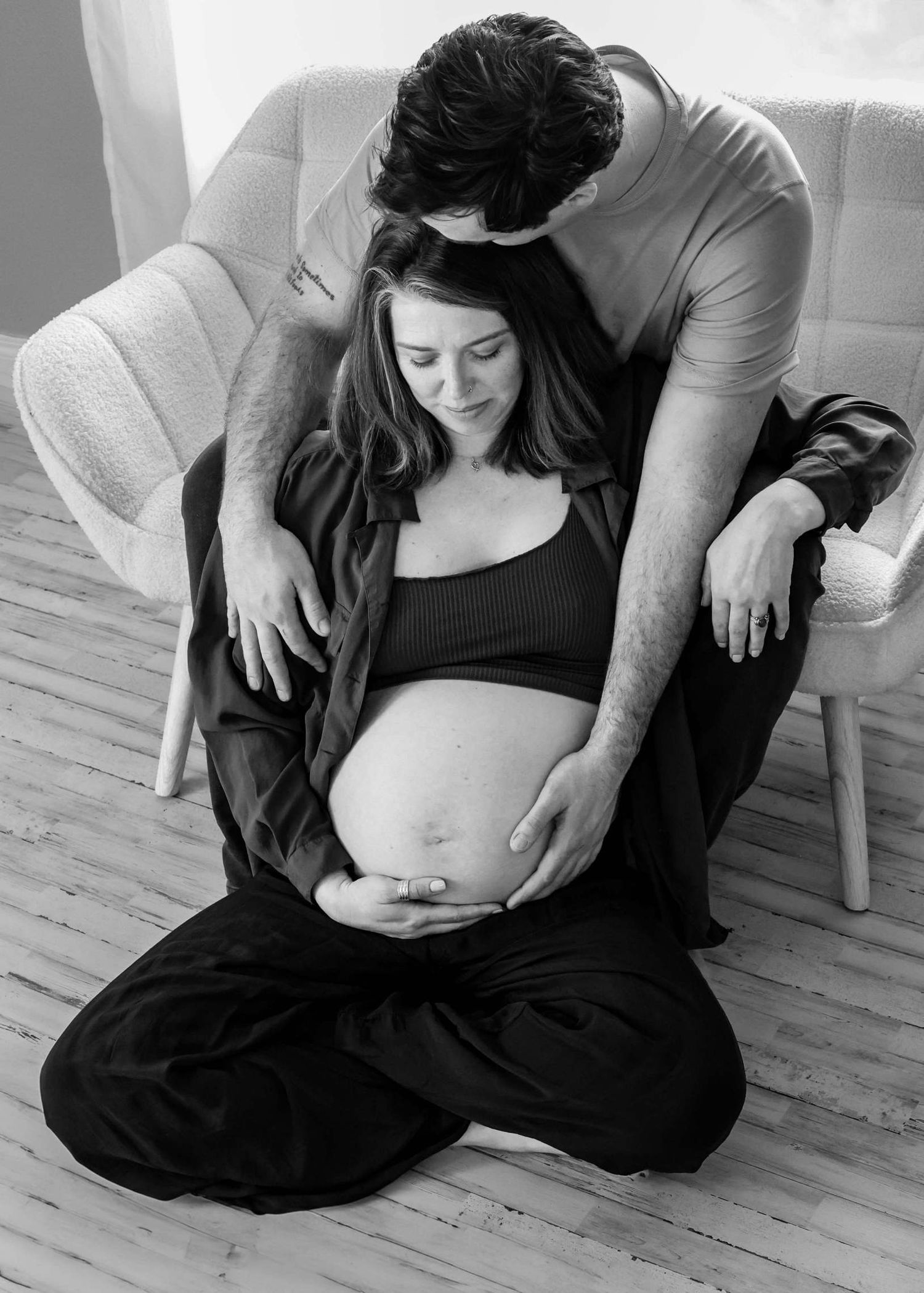 Black and white intimate maternity photo showing a couple embracing on a hardwood floor during a pregnancy photoshoot.
