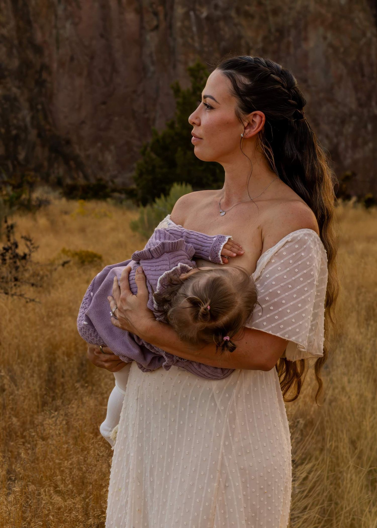 A woman in a white dress holds and embraces a small baby in a purple outfit against a golden field backdrop.