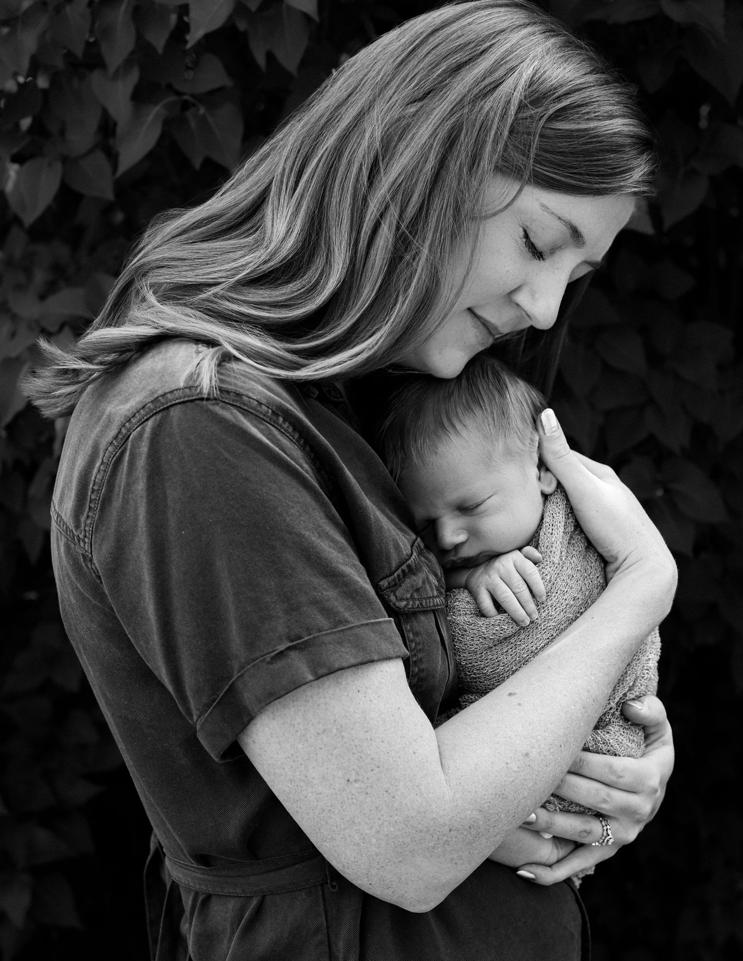 Black and white photo of a mother tenderly cradling her sleeping newborn baby close to her chest outdoors.