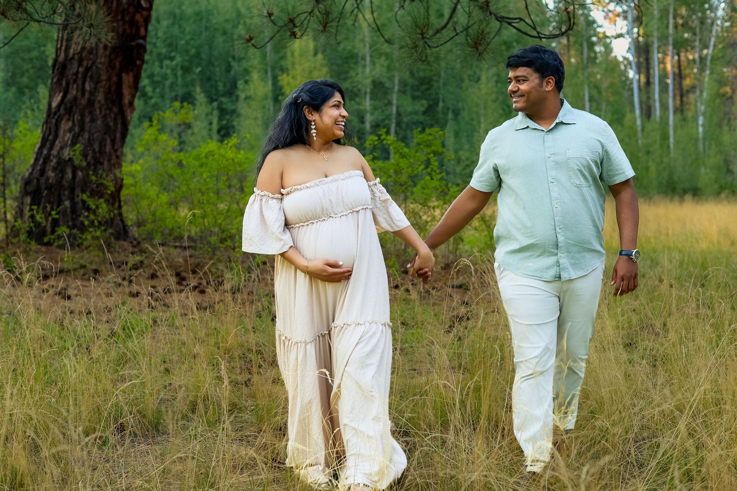 Pregnant woman in white dress holds hands with man in mint shirt, walking through a scenic forest clearing.