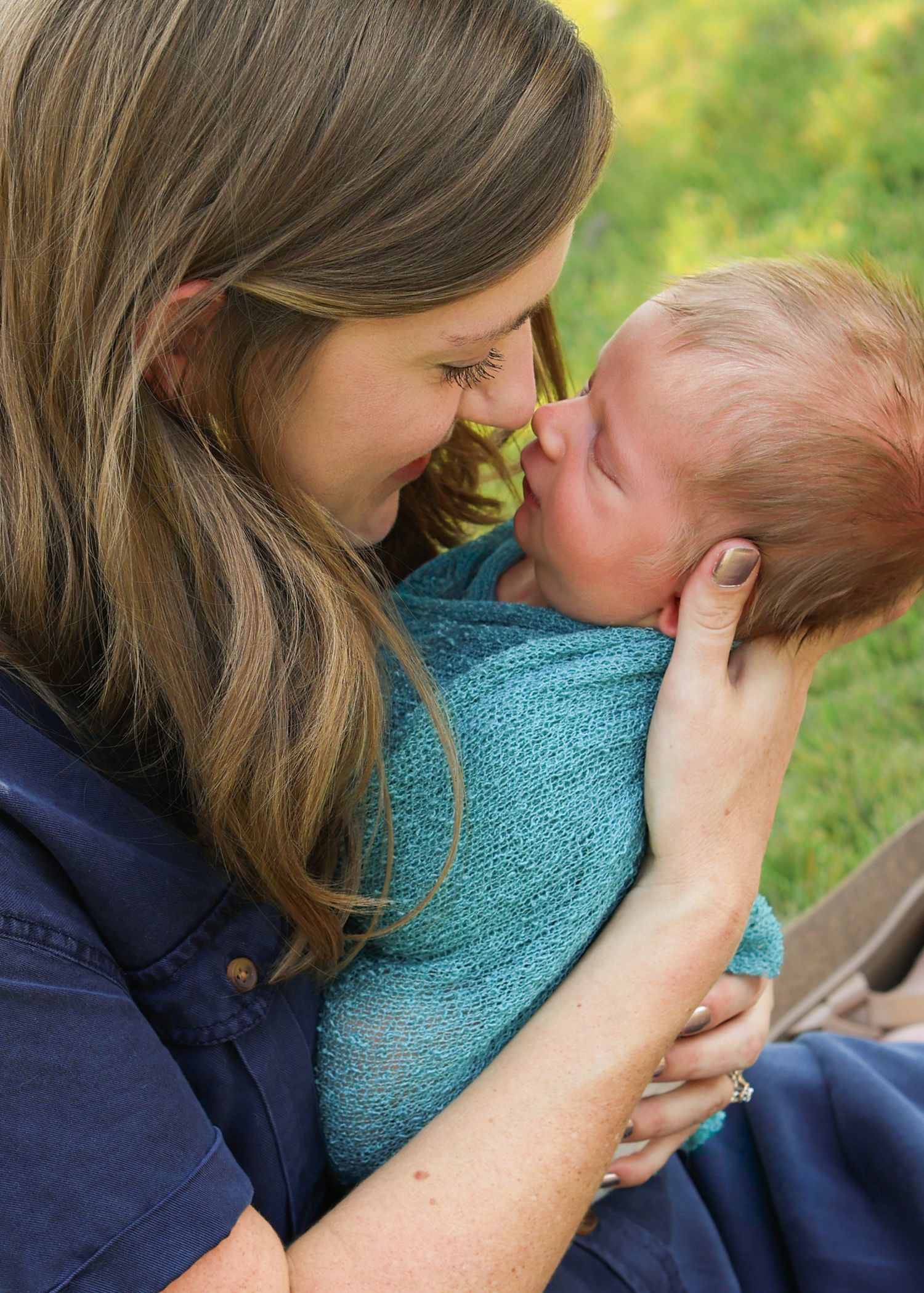 A mother tenderly touches noses with her newborn baby wrapped in a teal blanket outdoors.