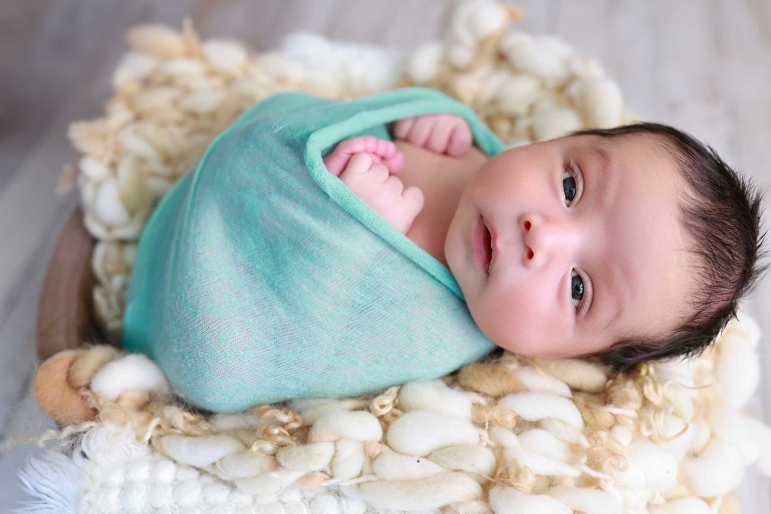 Smiling baby swaddled in mint green blanket lying on white textured backdrop.
