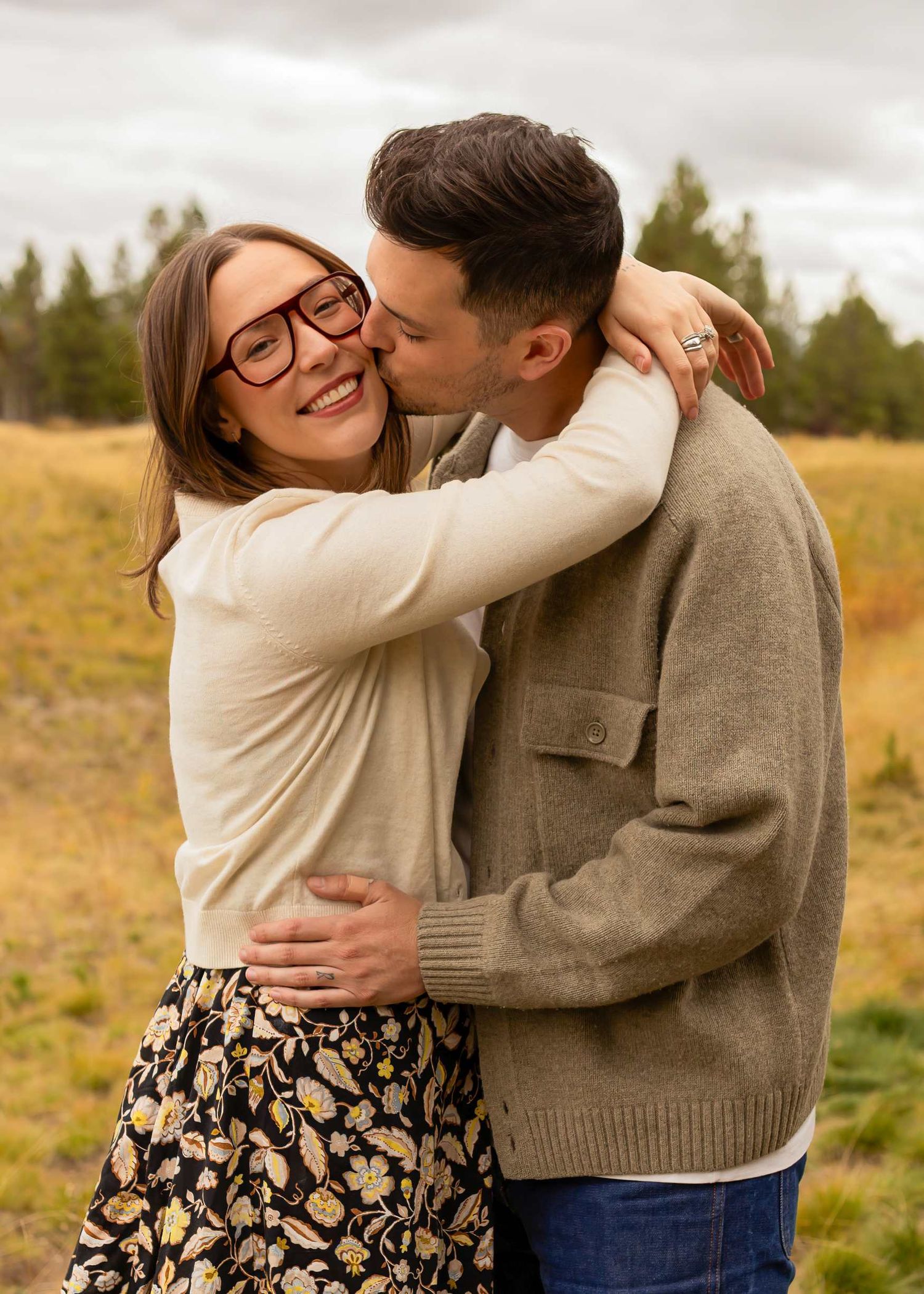 A couple embraces lovingly in a golden autumn field wearing casual sweaters and a floral skirt.