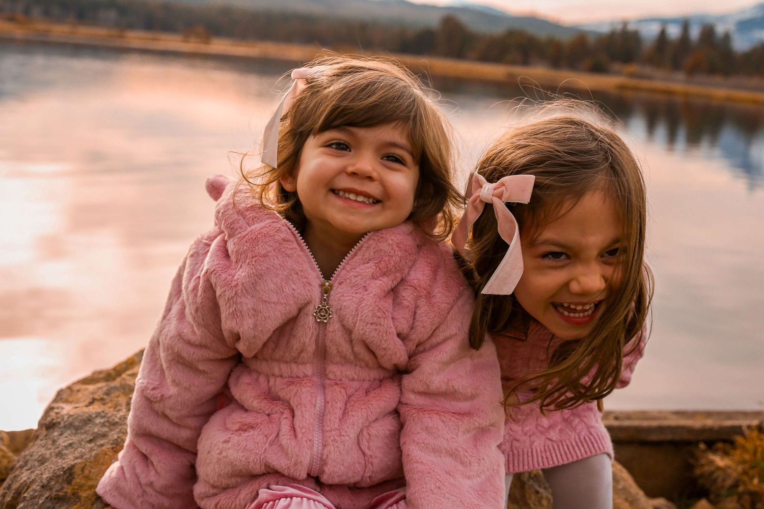 Two children in pink coats laugh and play together by a lake at sunset.