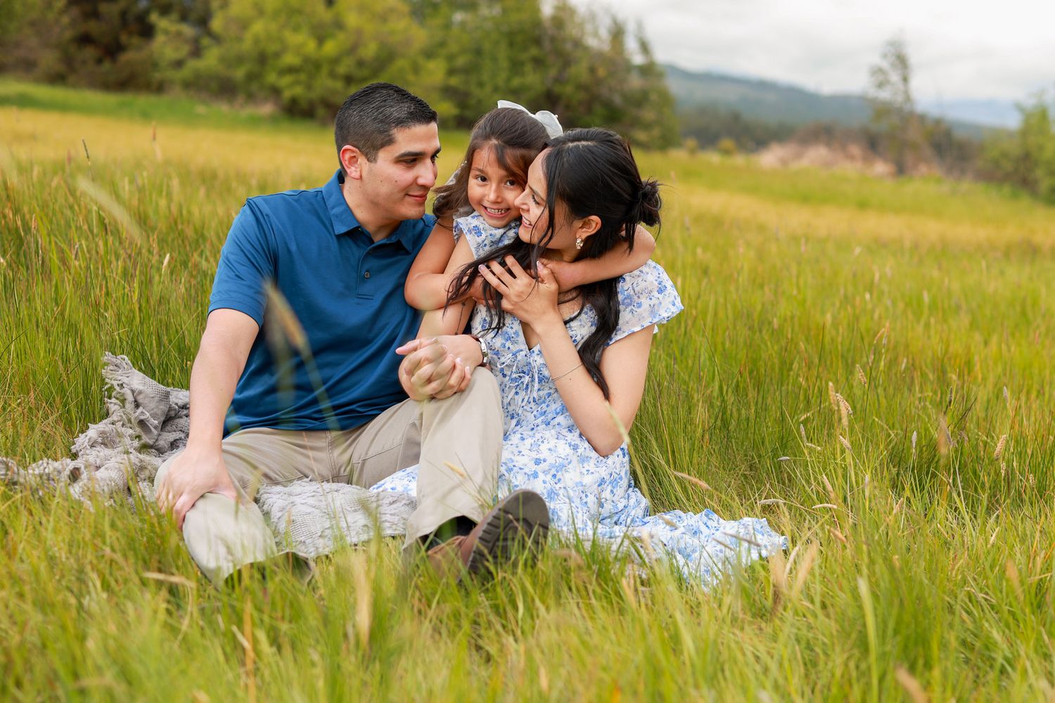 Family of three sitting in a green meadow, parents lovingly embracing their young daughter outdoors.