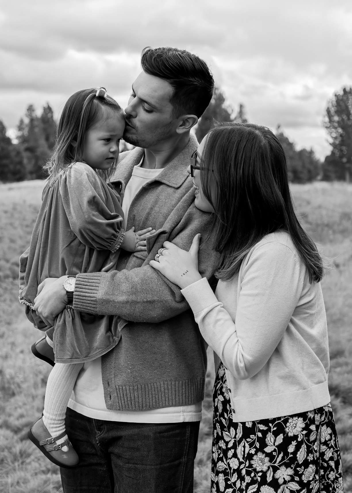 Black and white family portrait of parents kissing young daughter outdoors in a field with trees in the background. Sunriver Oregon