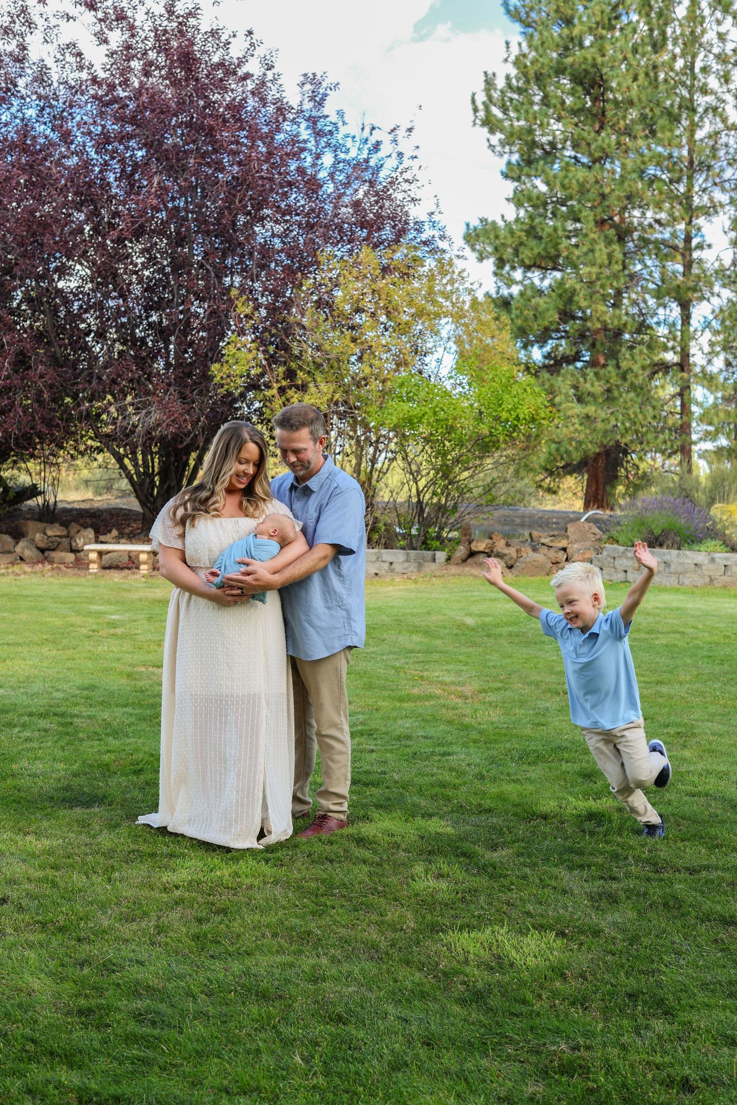 Family of four outdoors, parents cradling newborn while excited young boy jumps with arms raised on green lawn.