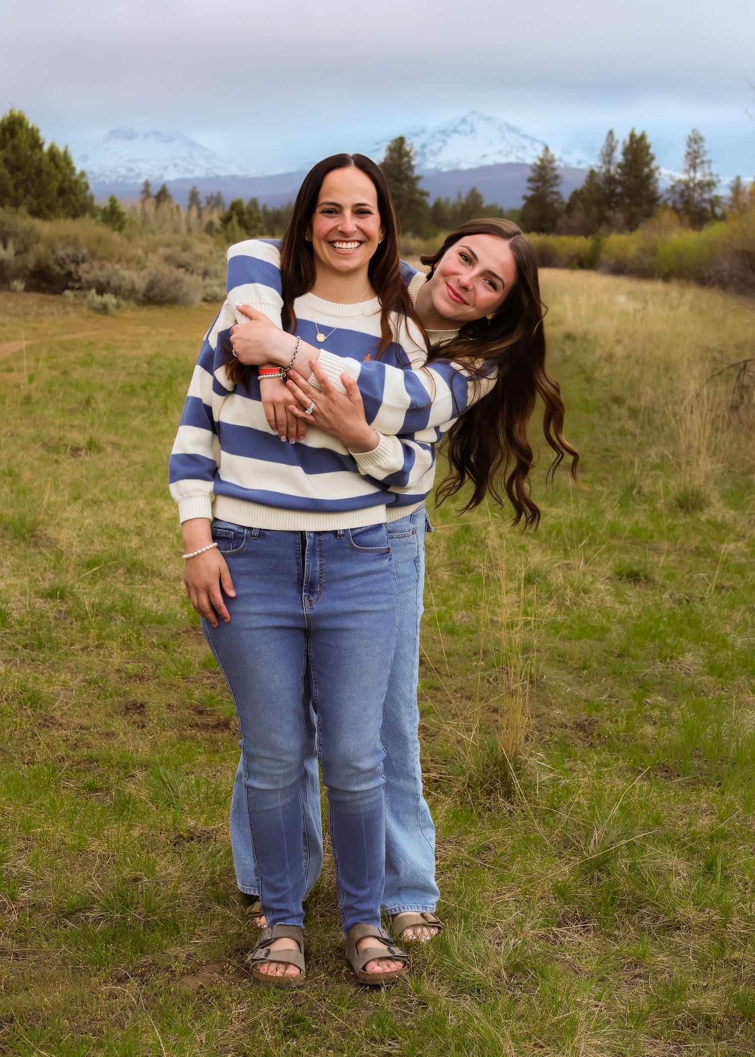 Two women smiling and posing together in a green field with snow-capped mountains in the background.