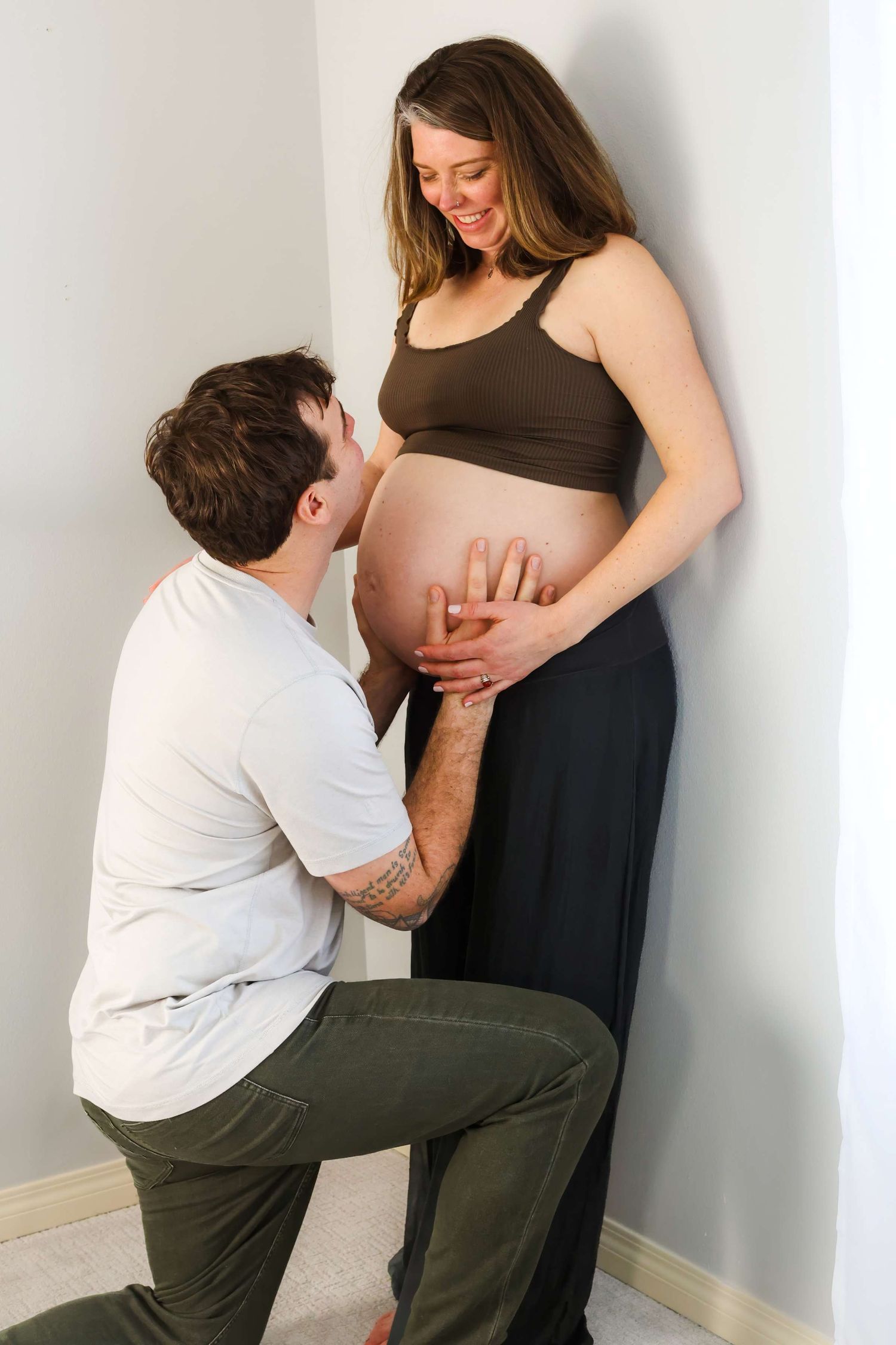 Man kneeling and touching pregnant woman's belly while she smiles, indoor maternity photo.