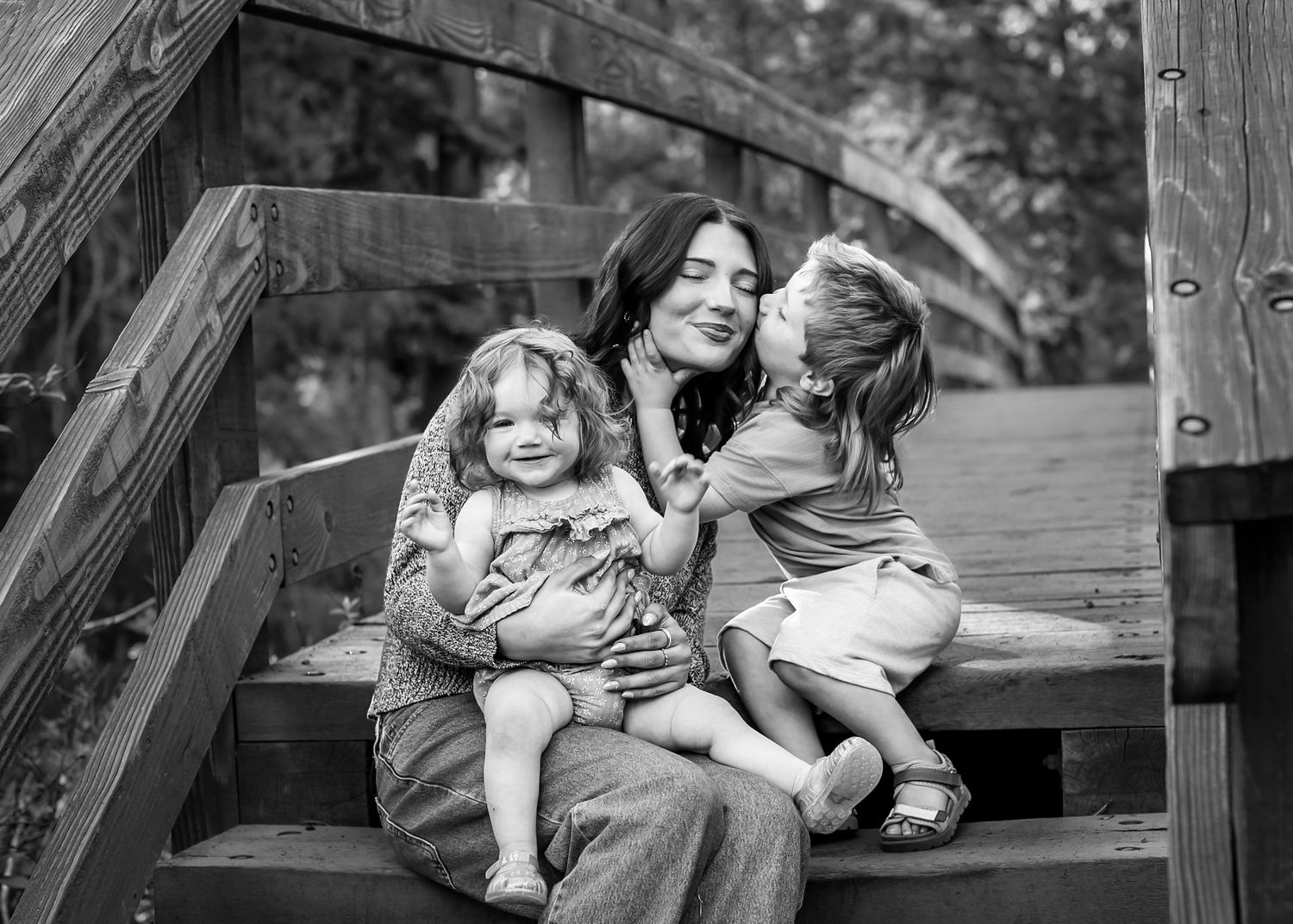 Black and white photo of a mother sitting on wooden steps with two young children hugging and kissing her cheeks. Shevlin Park Bend Oregon
