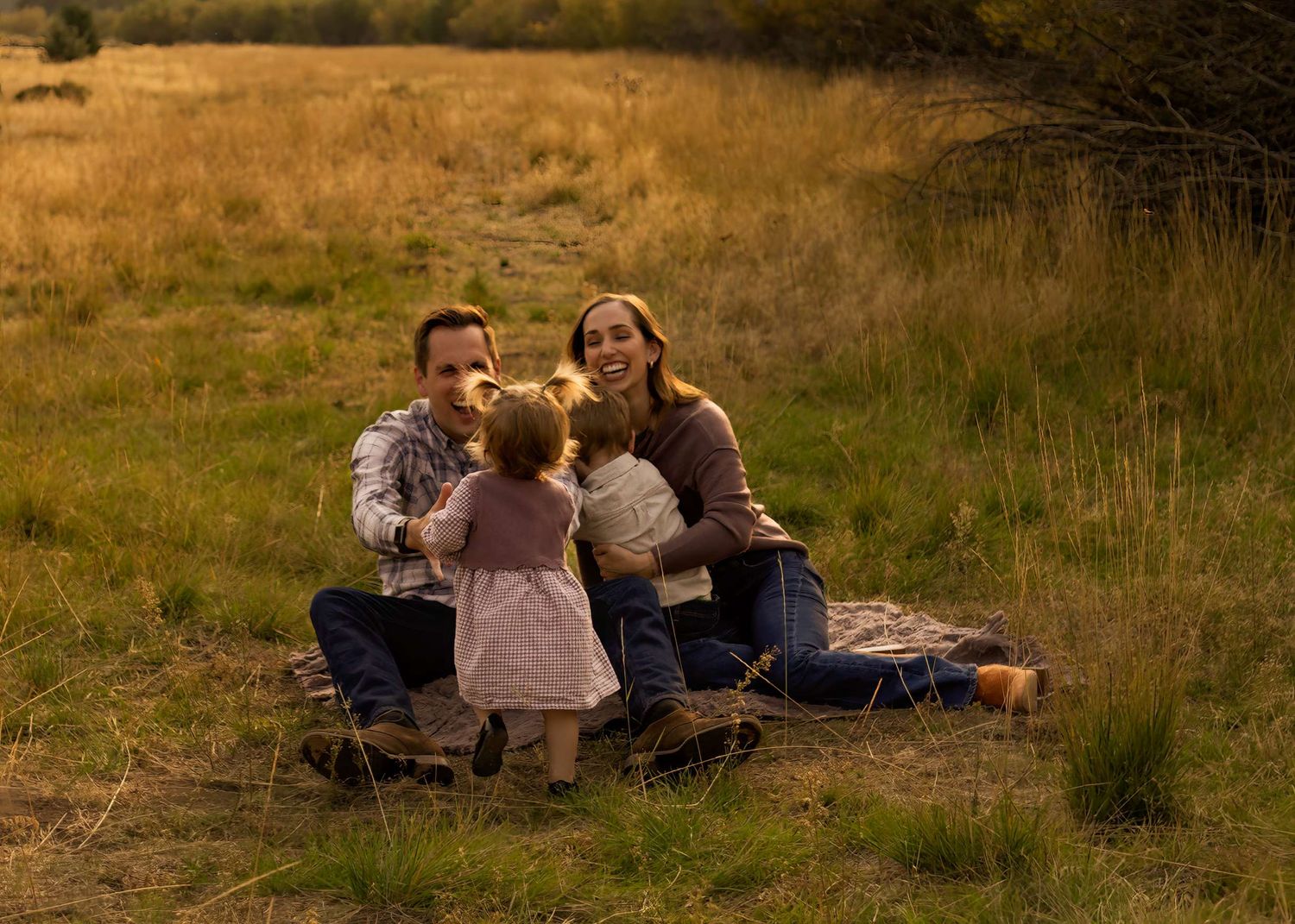Family sitting in golden meadow, parents smiling at toddler in pink dress holding treats outdoors. Indian Ford Meadows, Sisters Oregon