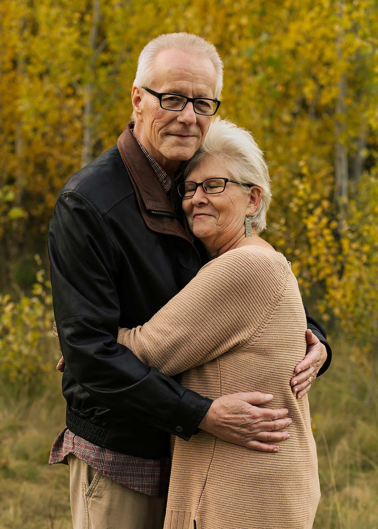 Elderly couple shares warm hugs and laughter in front of autumn trees.