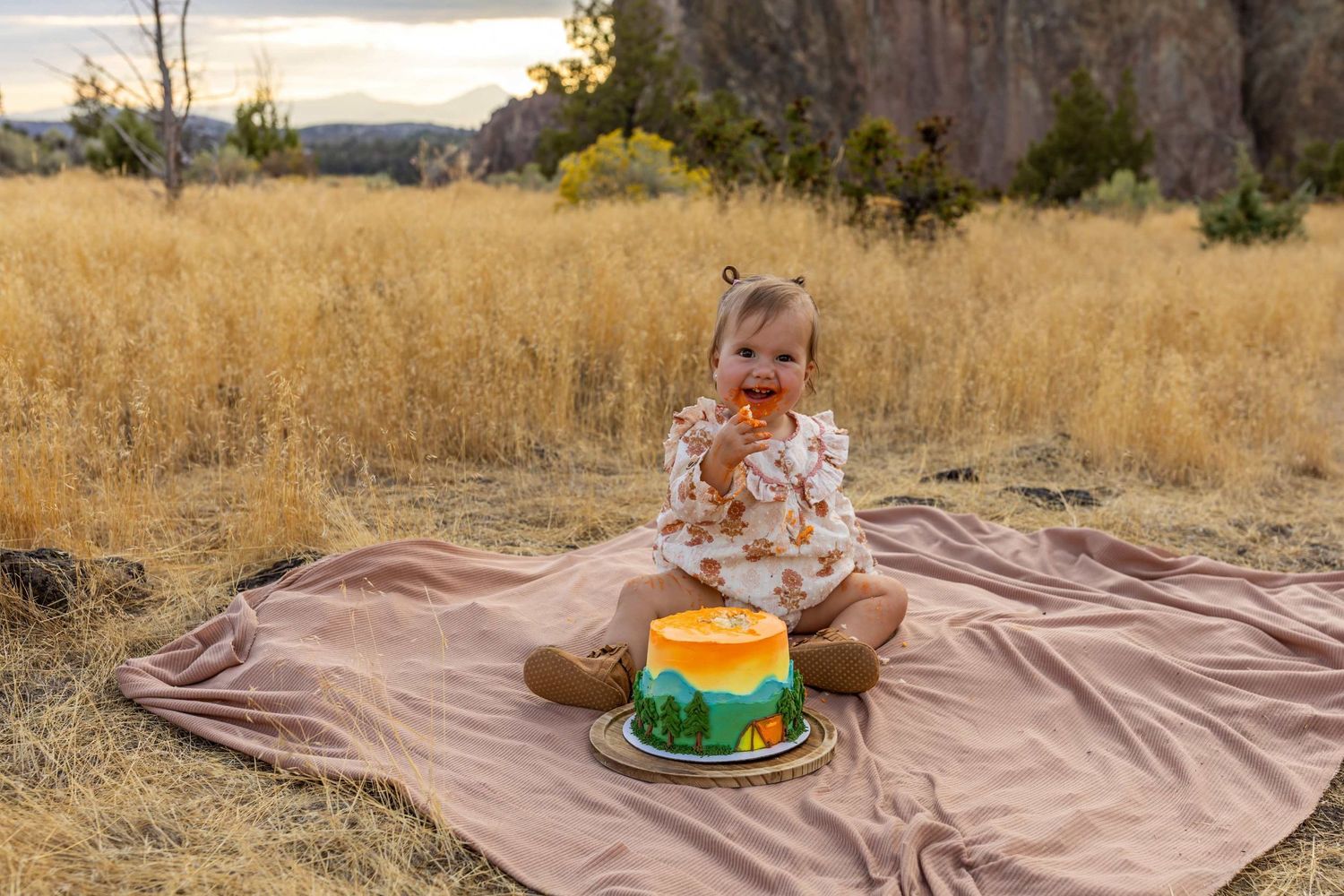First birthday cake smash photo session outdoors on brown blanket with autumn foliage in background.