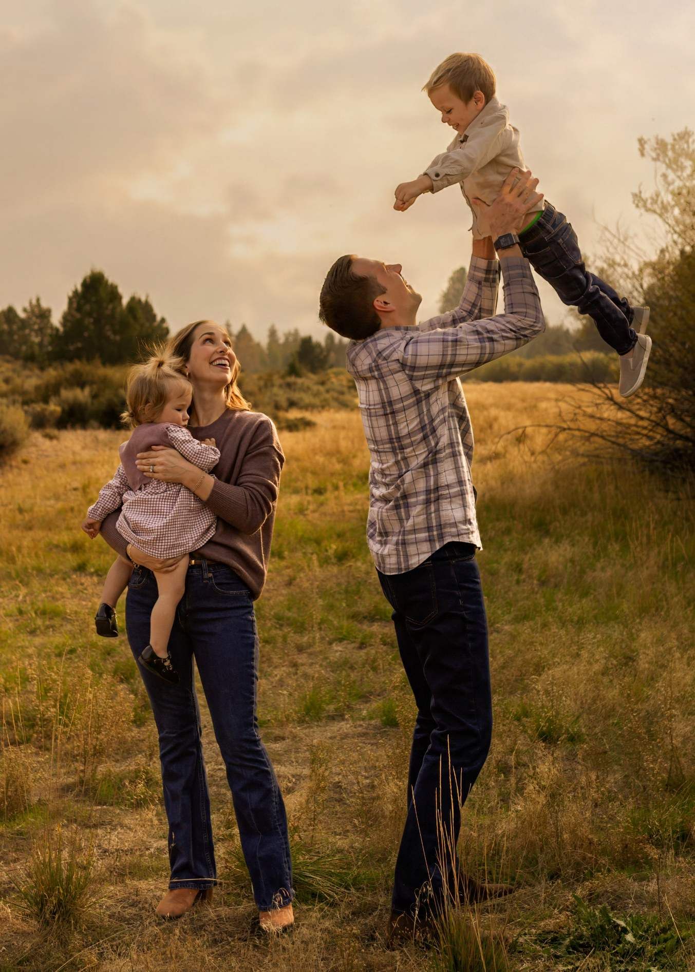 A couple shares a playful moment in a golden field at sunset, lifting and embracing each other with joy.