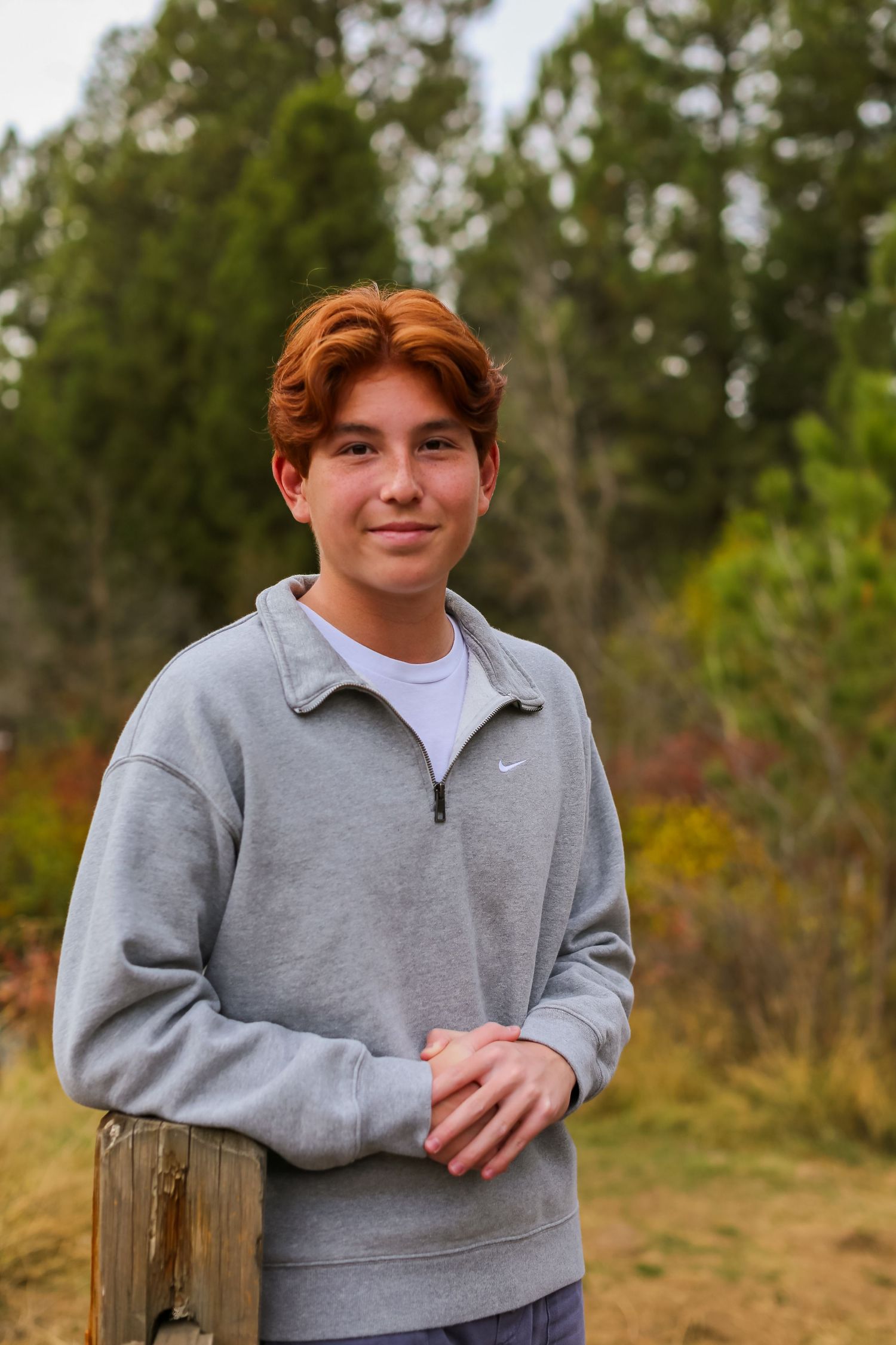 Teen boy with red hair wearing a gray Nike quarter-zip sweatshirt standing outdoors near trees.