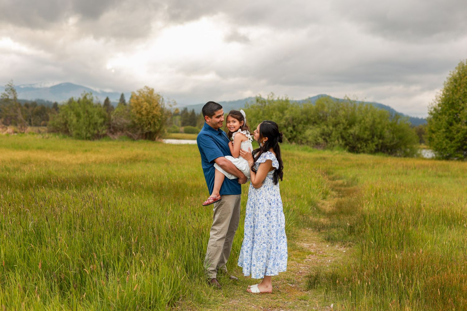 Family of three standing in a lush green meadow with mountain views and cloudy skies in the background.