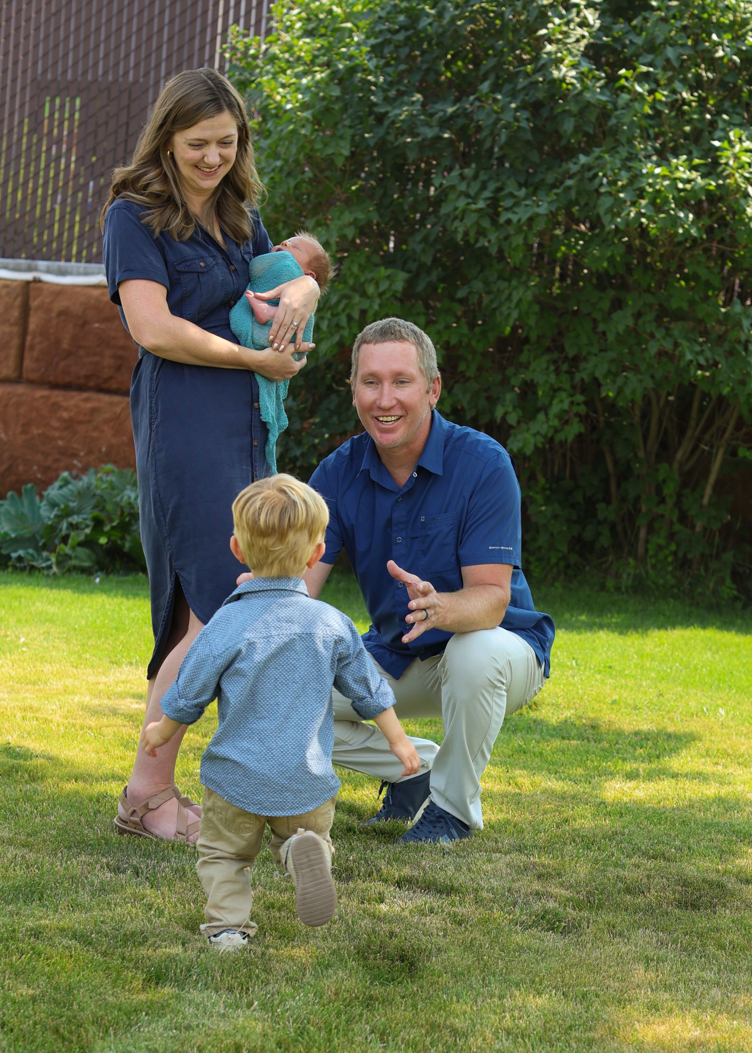 A happy family outdoors on green grass, with a woman holding a newborn and a man crouching to greet a toddler.