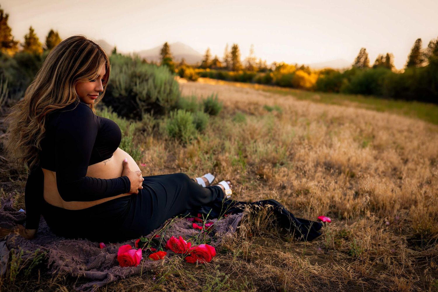 Pregnant woman in black outfit sits on blanket in golden field at sunset with red flowers nearby. Indian Ford Meadows, Sisters Oregon