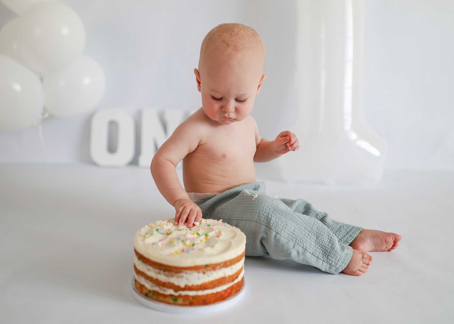 A baby sits next to a white frosted cake on a wooden board during a cake smash photo session.