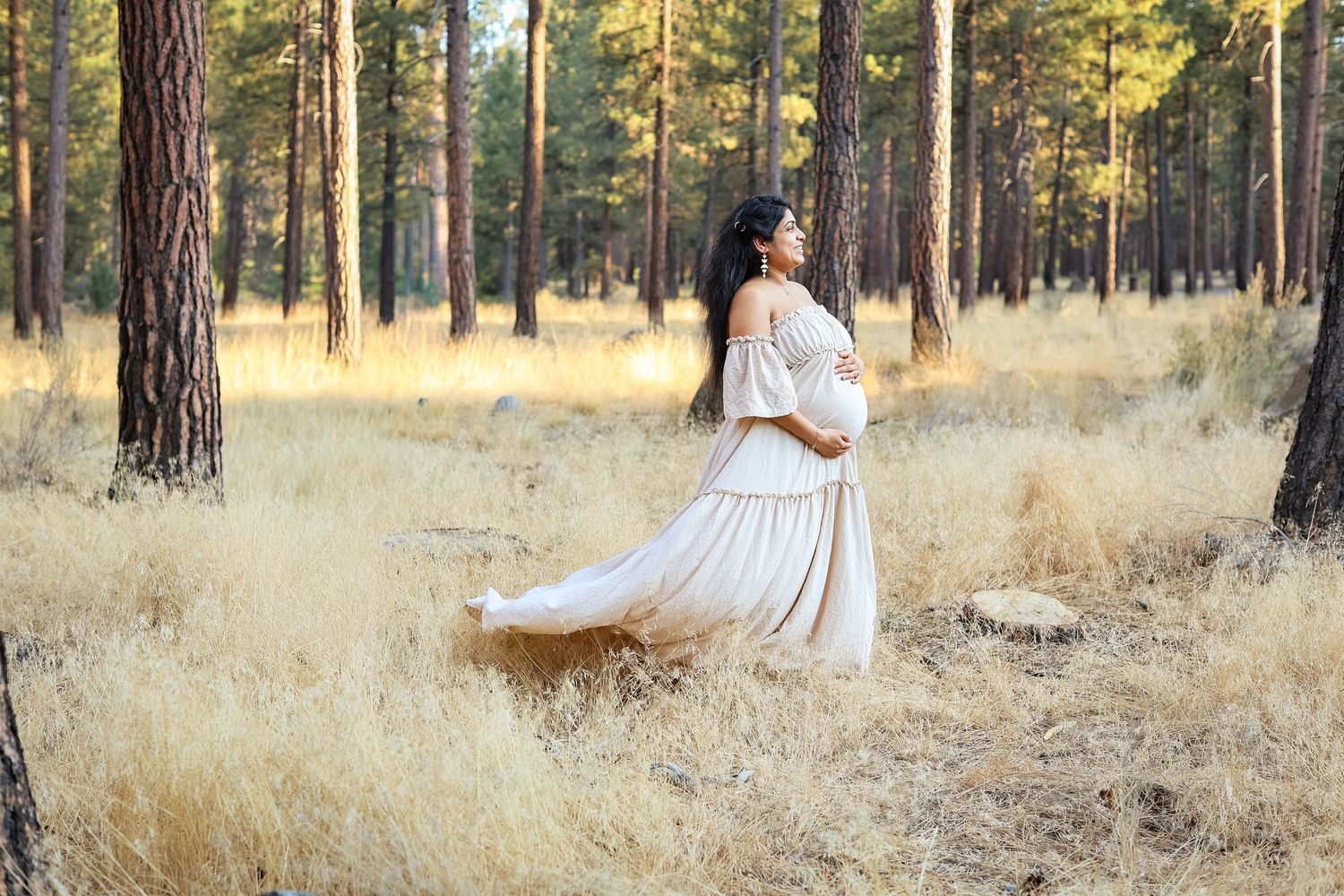 Pregnant woman in flowing white off-shoulder gown standing in sunlit pine forest with golden grass.