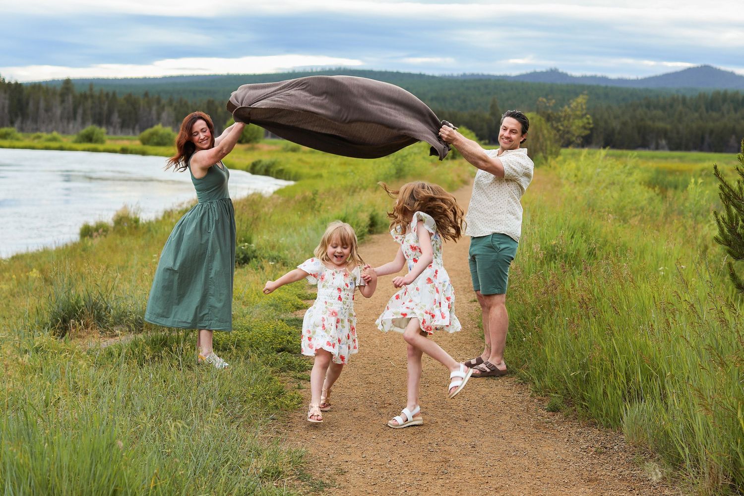 Family of four playing outdoors near a river, parents waving a blanket over two young girls on a scenic trail.