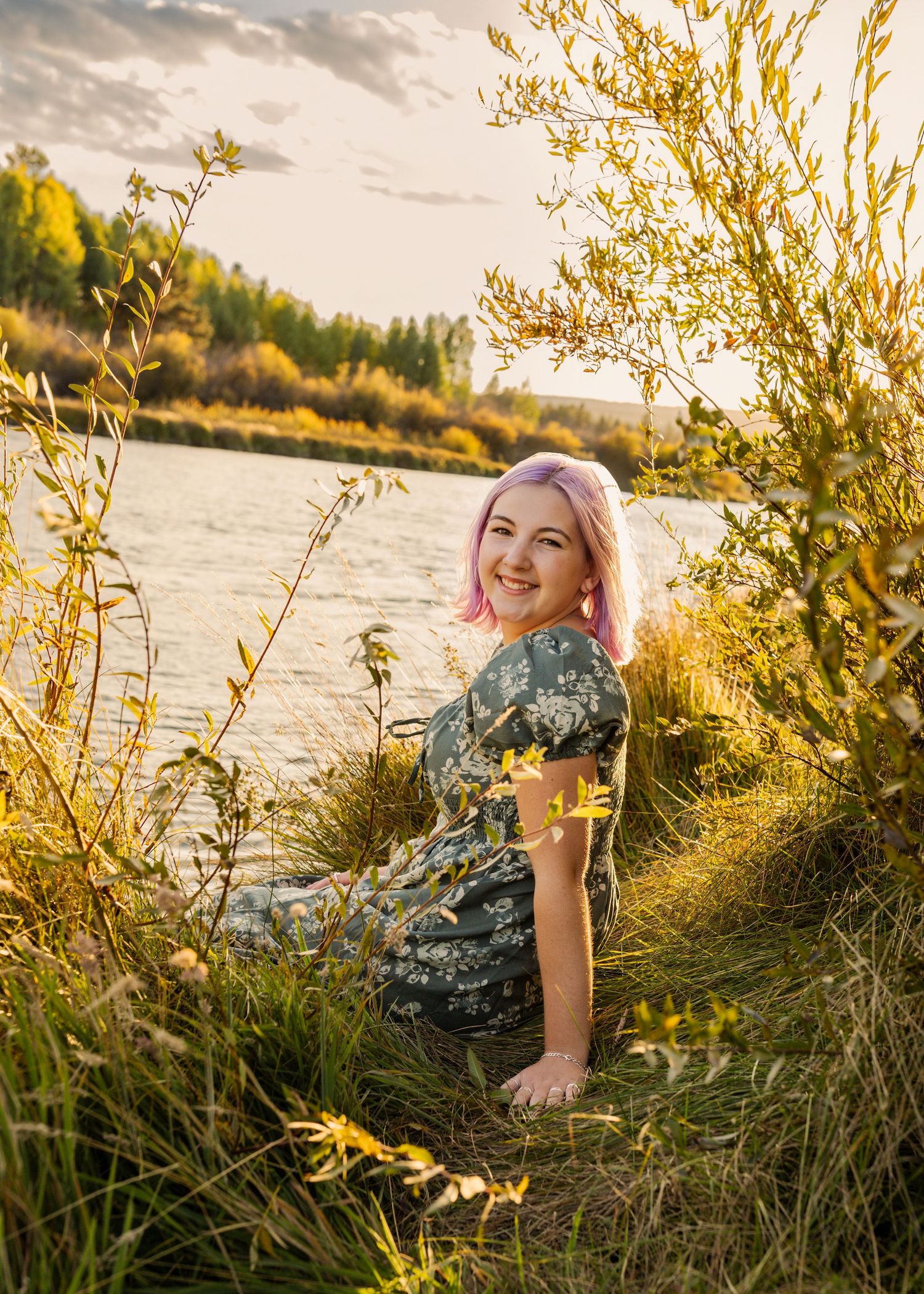 Woman with pink hair in floral dress sitting by a sunlit river surrounded by golden wildflowers at sunset.