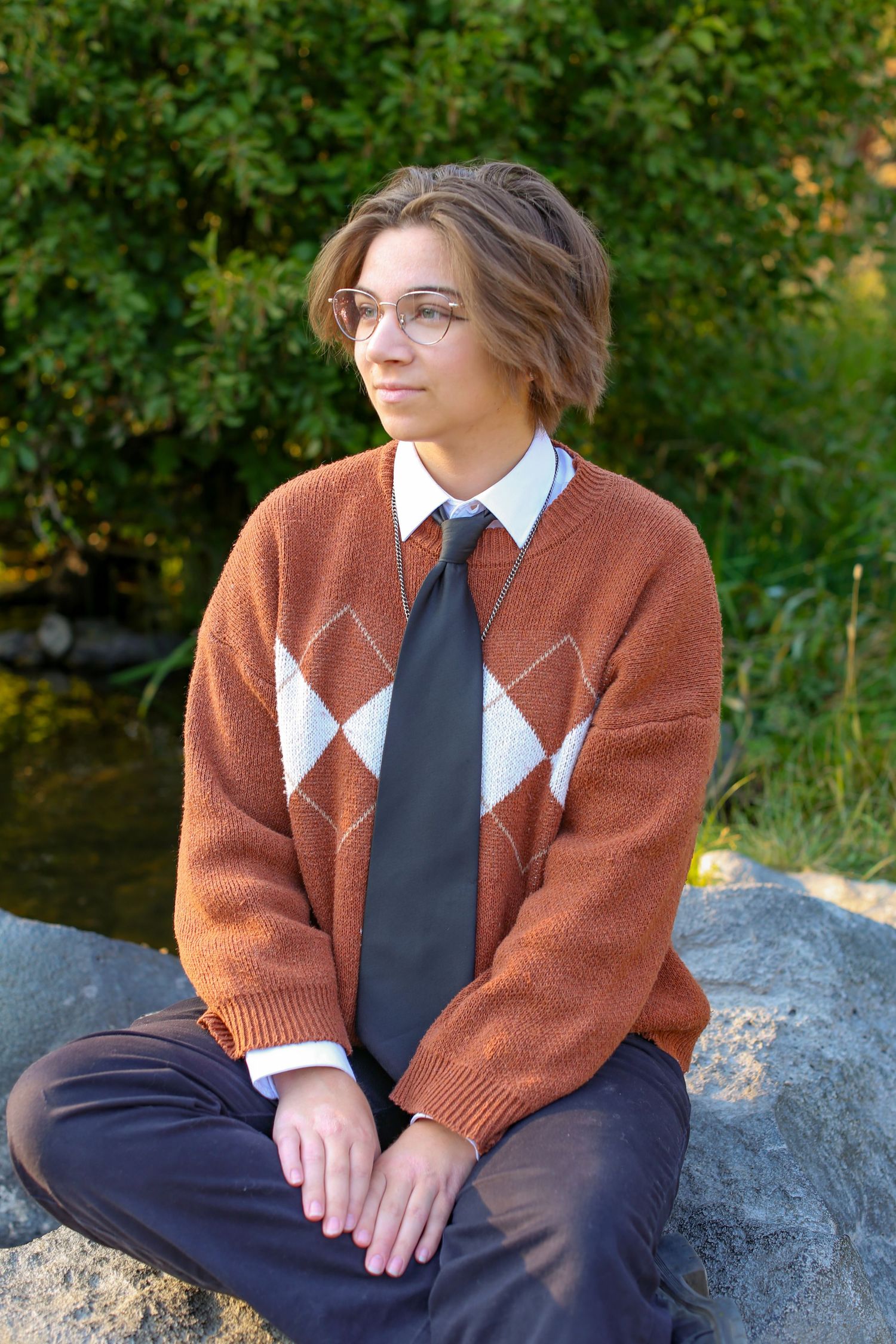 Young person in argyle sweater, tie, and glasses sitting on rocks outdoors near greenery.