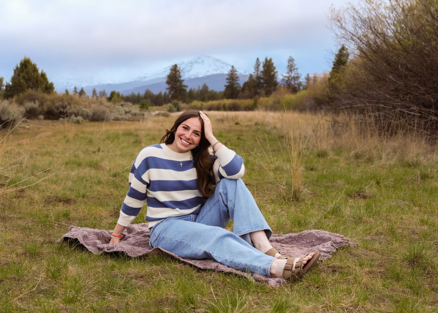 Young woman in striped sweater and jeans sitting on blanket in meadow with snow-capped mountain backdrop.