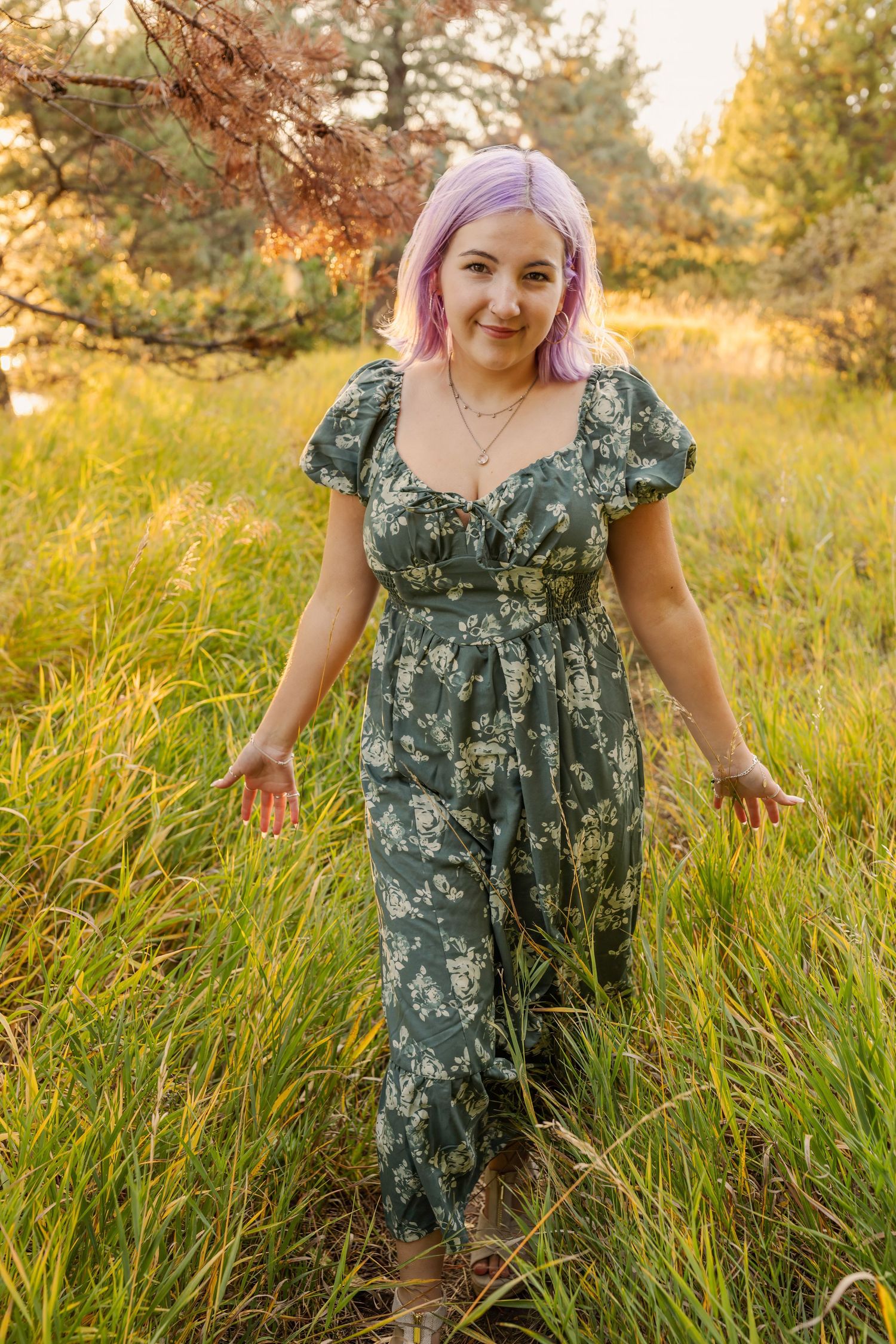 Woman with pink hair wearing a green floral jumpsuit standing in tall grass during golden hour.