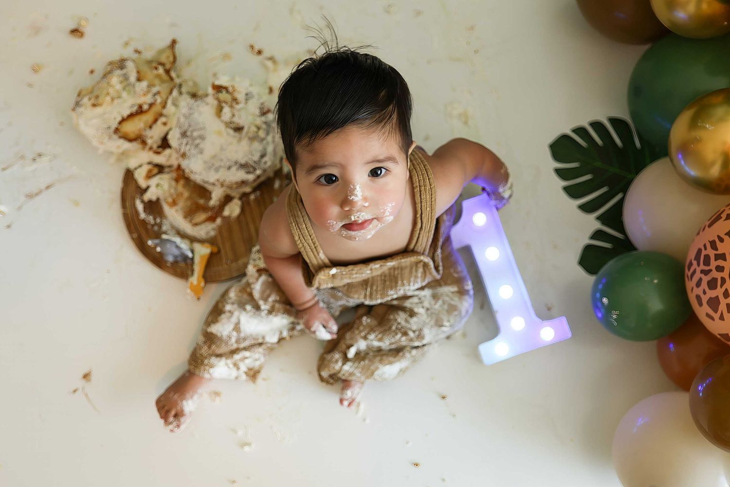 A small child lies on the floor surrounded by scattered cake crumbs and frosting during a cake smash photo session.