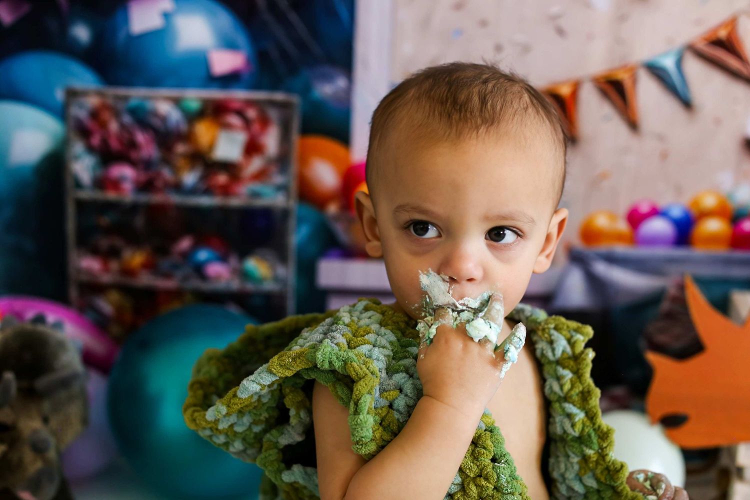 Baby wearing leafy green garland enjoys cake during colorful birthday celebration.